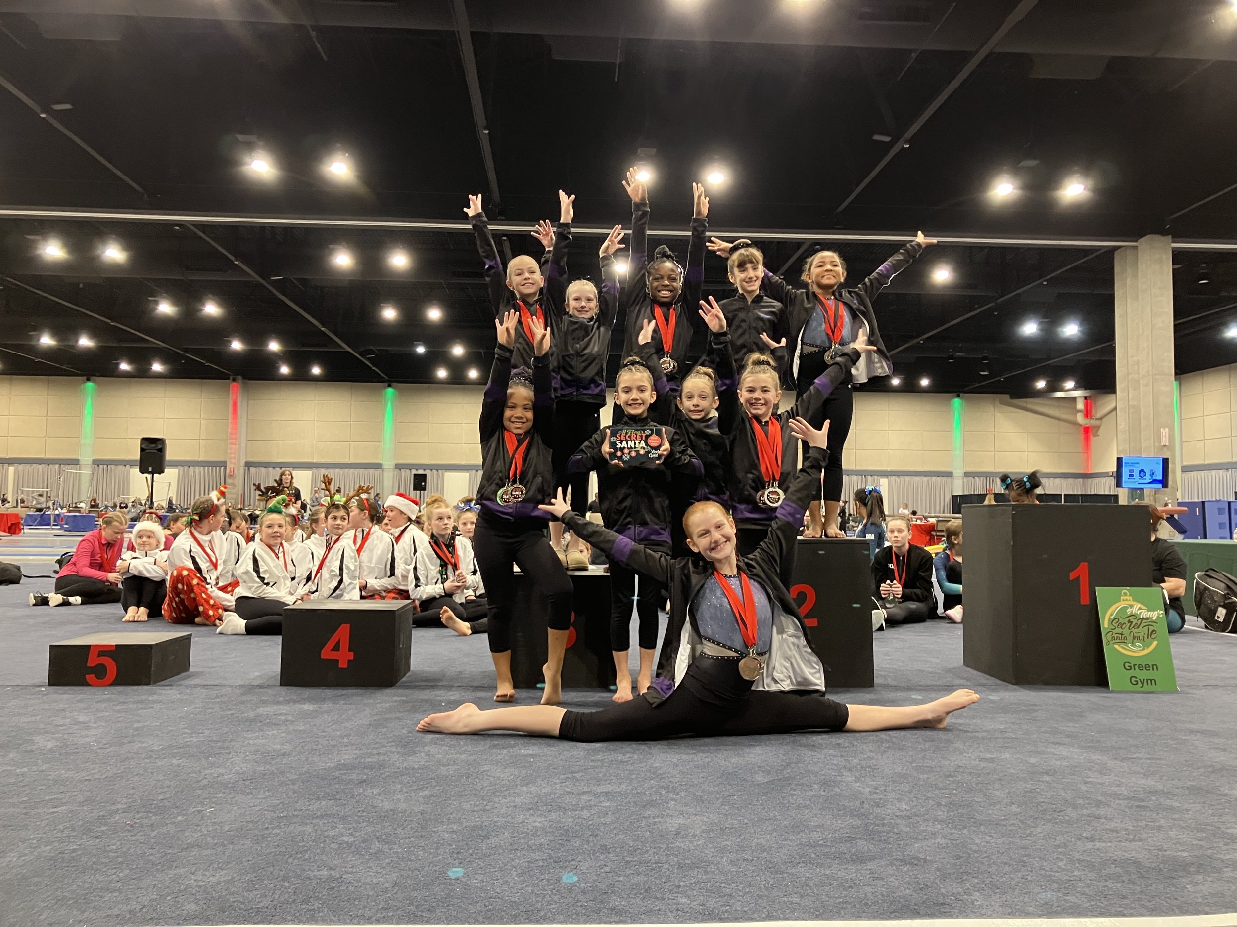 A group of young gymnasts standing on a podium celebrating, with some making victory signs, in a gymnasium decorated for a holiday event, with children sitting on the floor in the background.