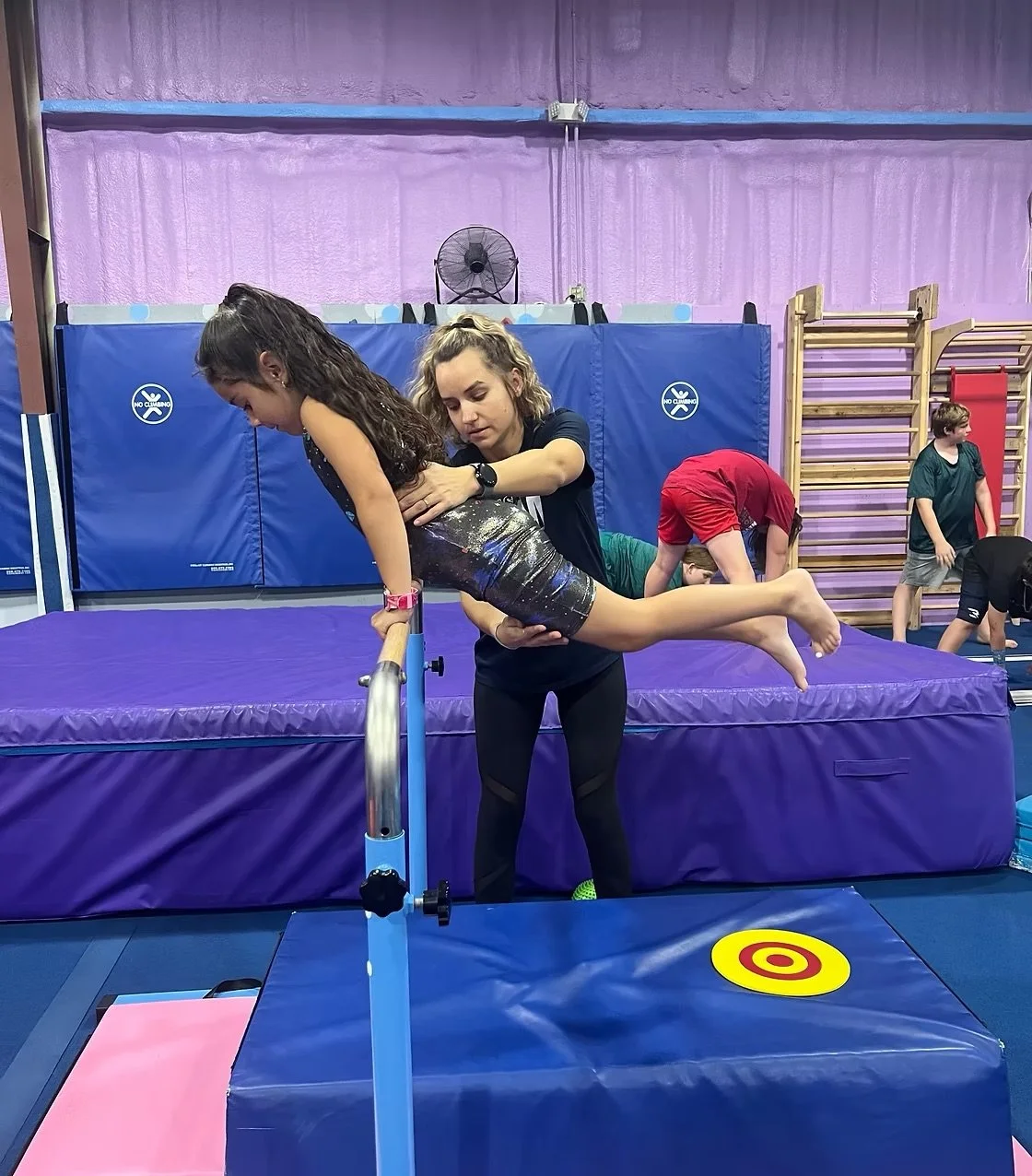A young girl practicing hand balancing on a padded block at an indoor gym with her instructor supporting her from behind.