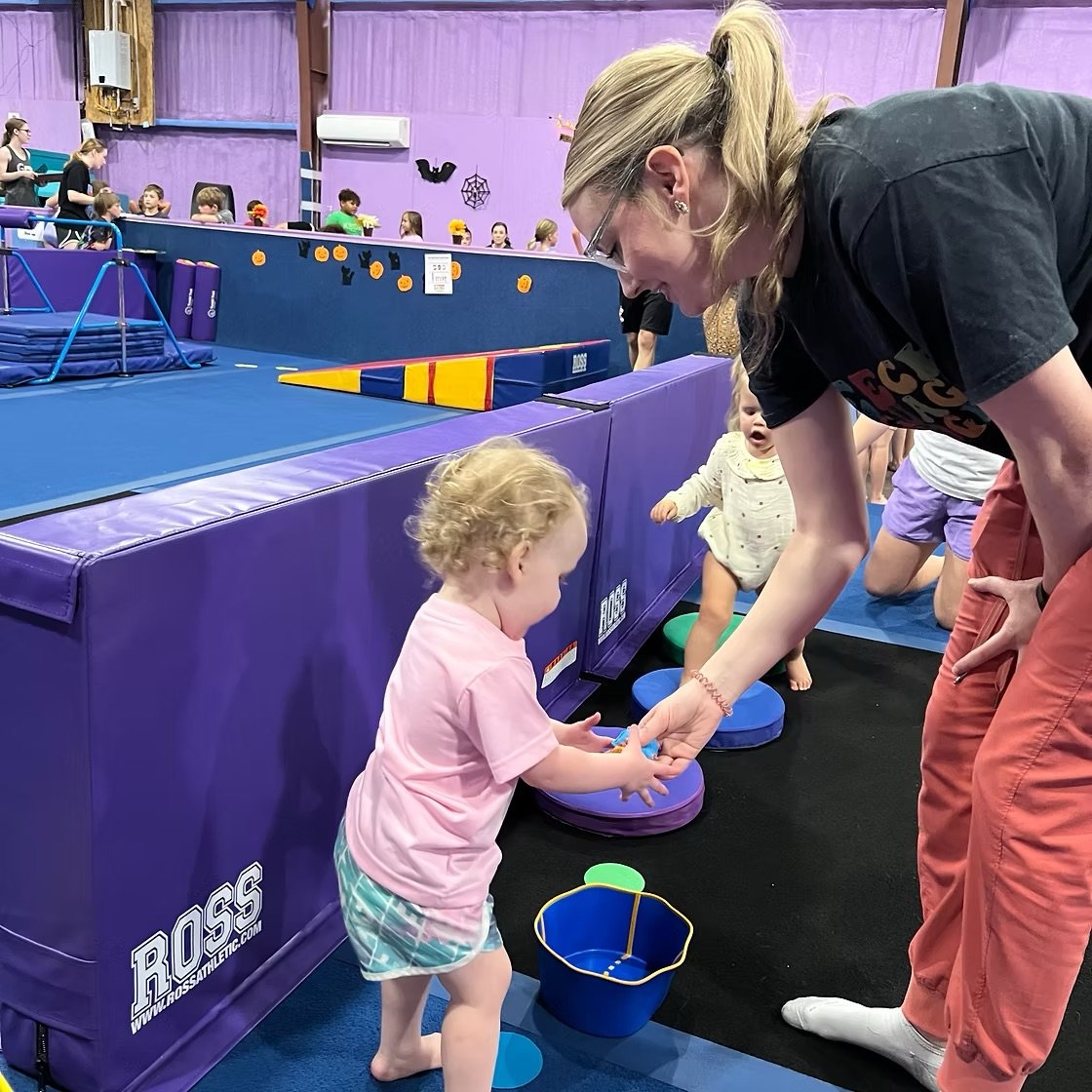 A woman helping a young child with a game or activity at an indoor play area with purple padding and various children in the background.