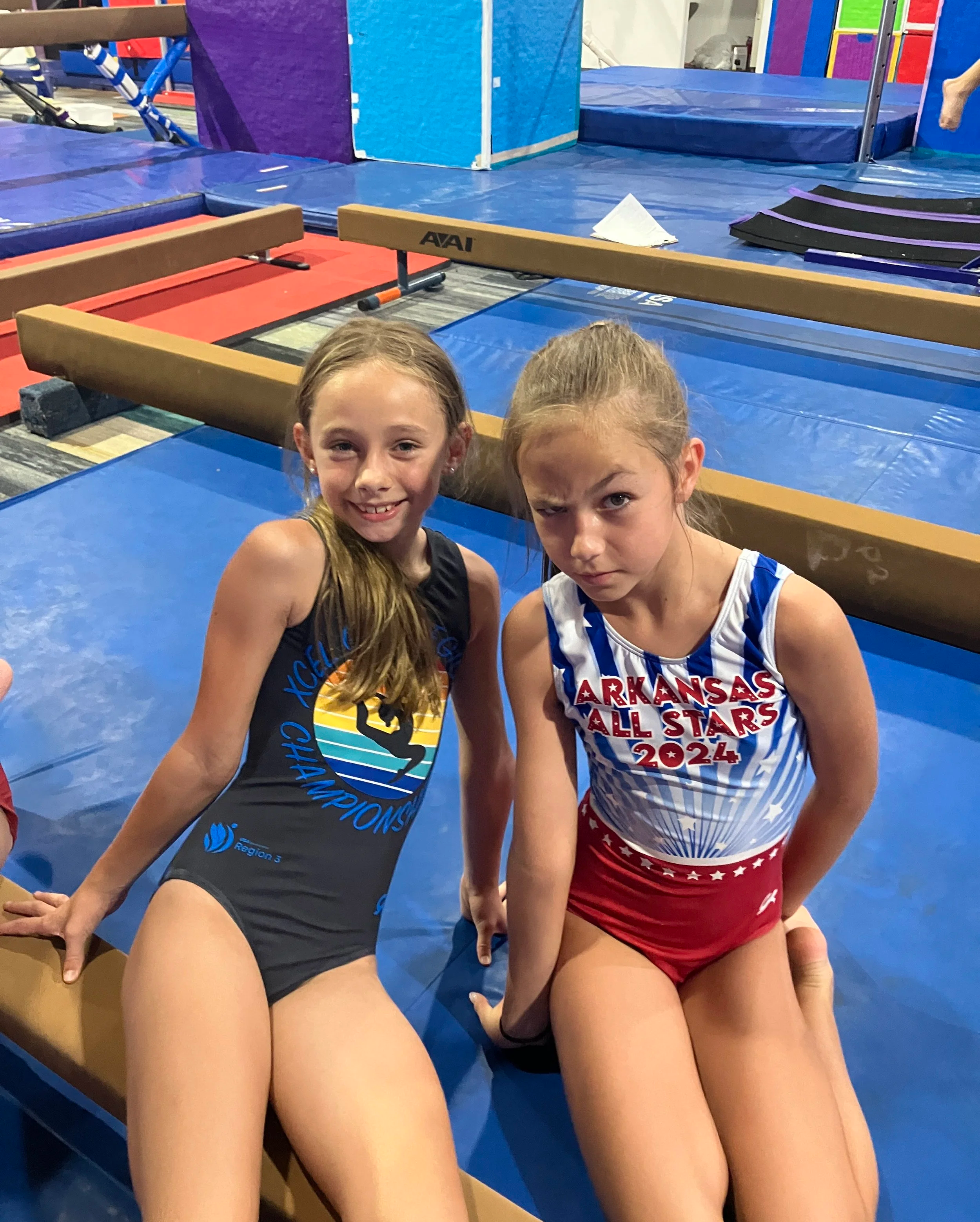 Two young female gymnasts in athletic outfits sitting on the floor of a gymnasium, with gymnastics equipment such as mats, beams, and a foam pit in the background.