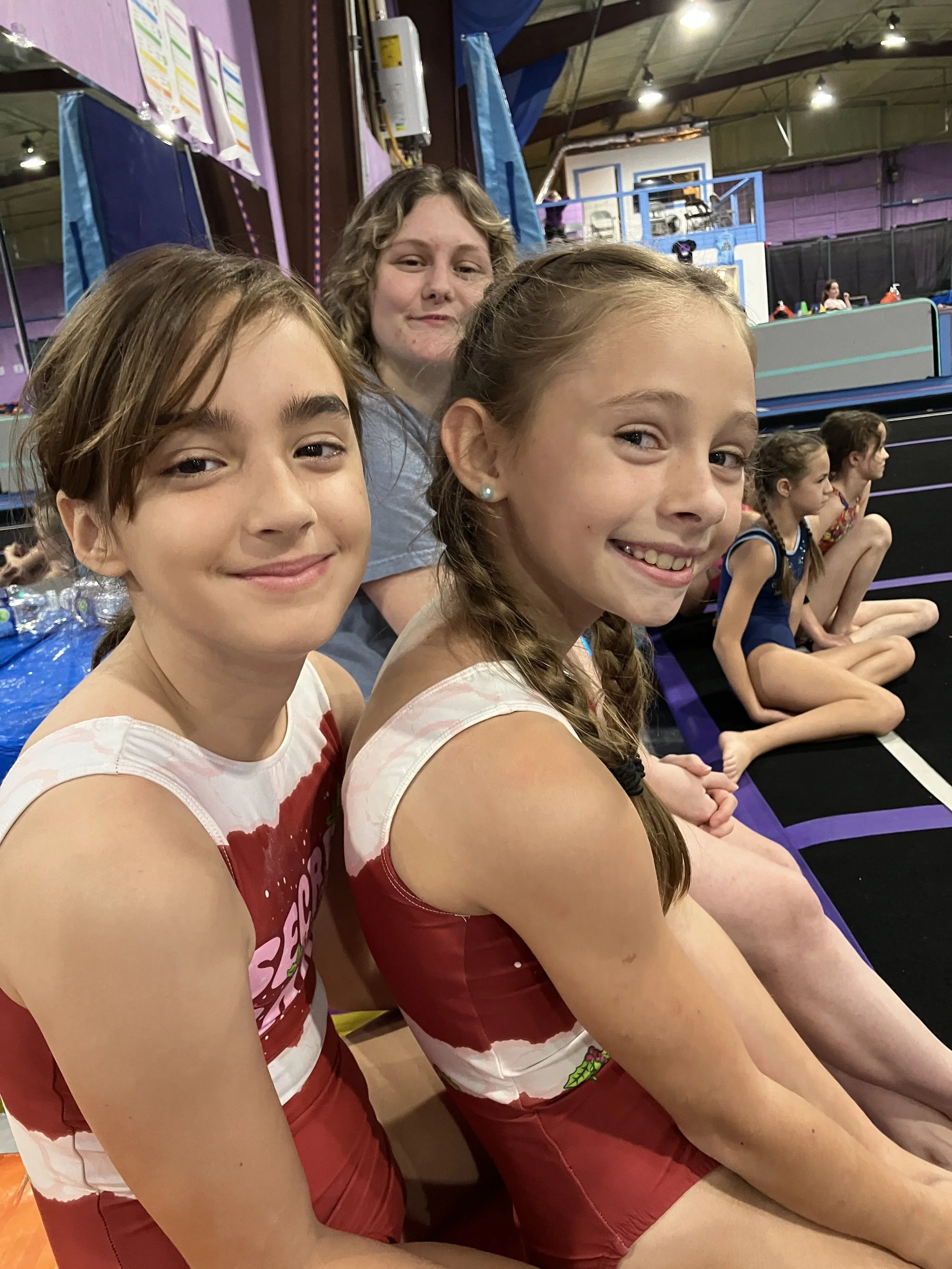 Three smiling young girls sitting in a row at an indoor trampoline park or recreational center, with a woman behind them, and others in the background.