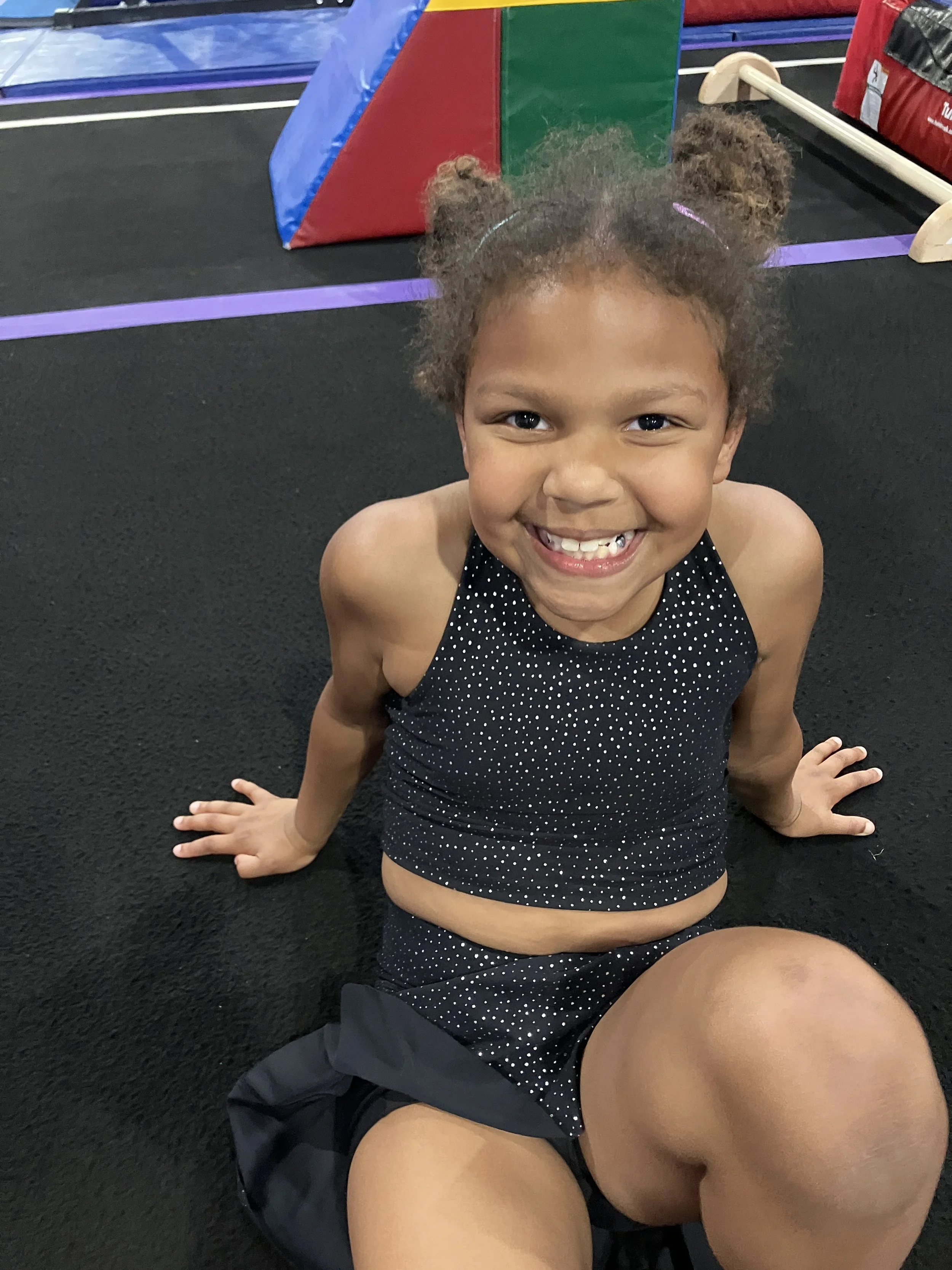 A smiling young girl with curly hair styled in puffs, wearing a black dress with white polka dots, sitting on a mat in an indoor gym area. Hot Springs Gymnastics | 112 Bluestone Ct. Hot Springs, Arkansas 71901