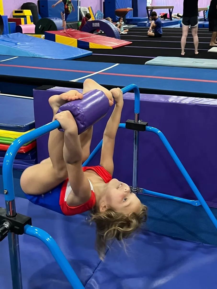 Child with long hair hanging upside down on a gymnastics bar in an indoor gym, surrounded by various gymnastic equipment and other children practicing in the background.