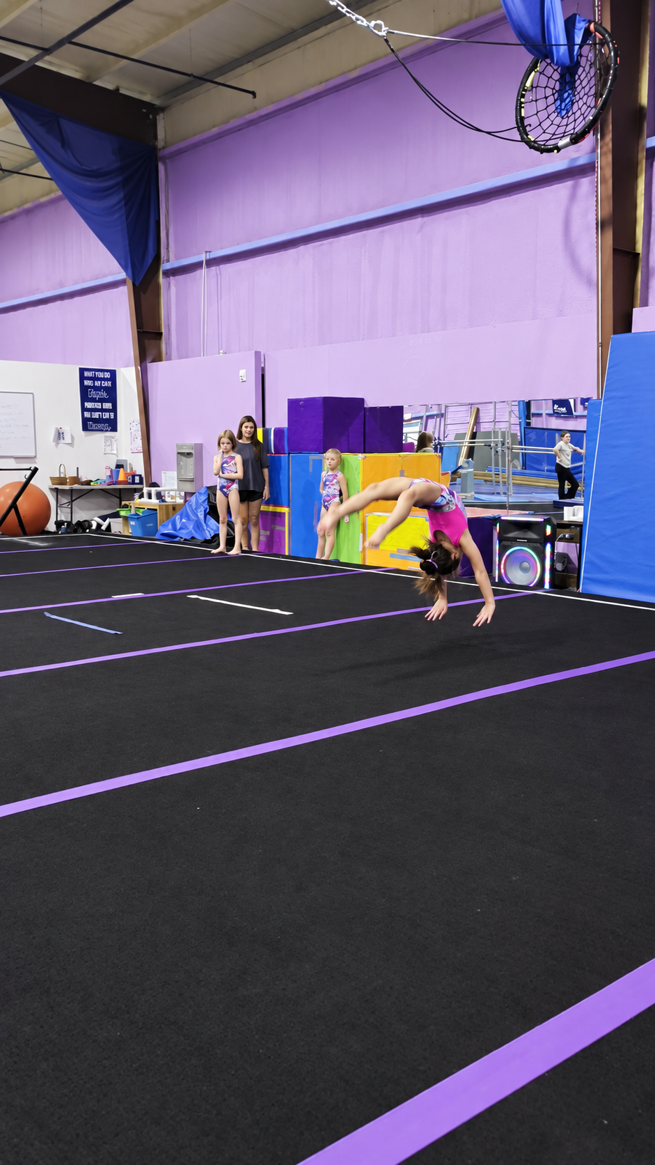A girl is performing a backflip on a gymnastics black mat with purple lines in an indoor gym, surrounded by colorful equipment and three girls watching.