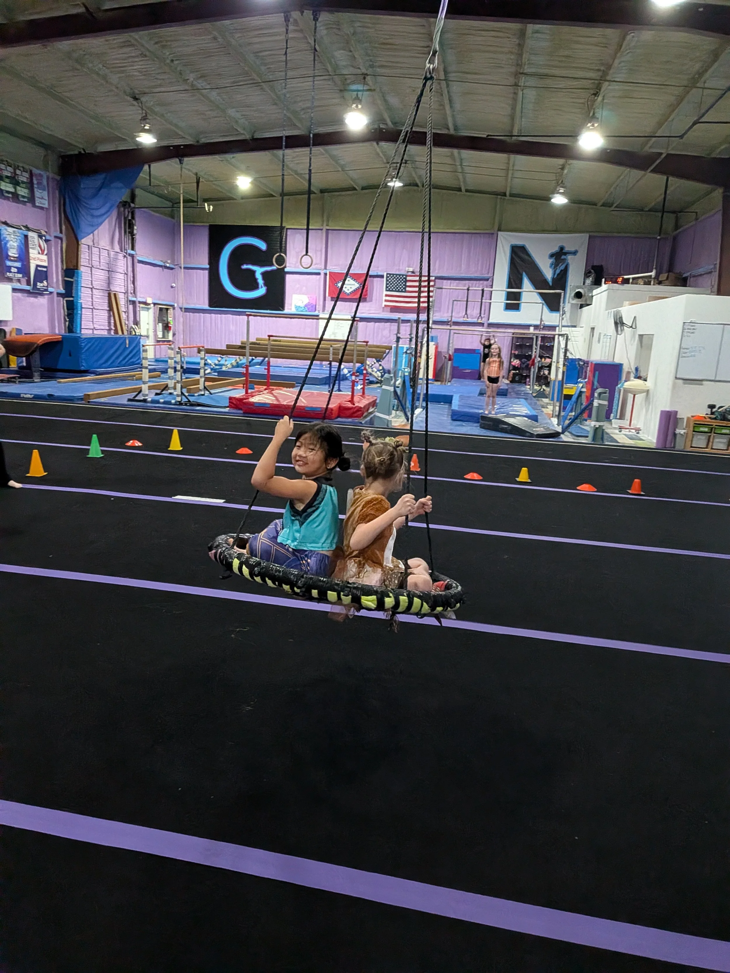 Children playing on gymnastic swings inside a gymnasium with purple walls, American flag, gymnastic equipment, and large banners on the walls.