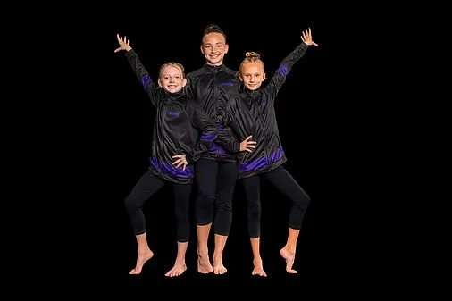 Three young female gymnasts in black and purple uniforms posing with arms raised in front of a black background.