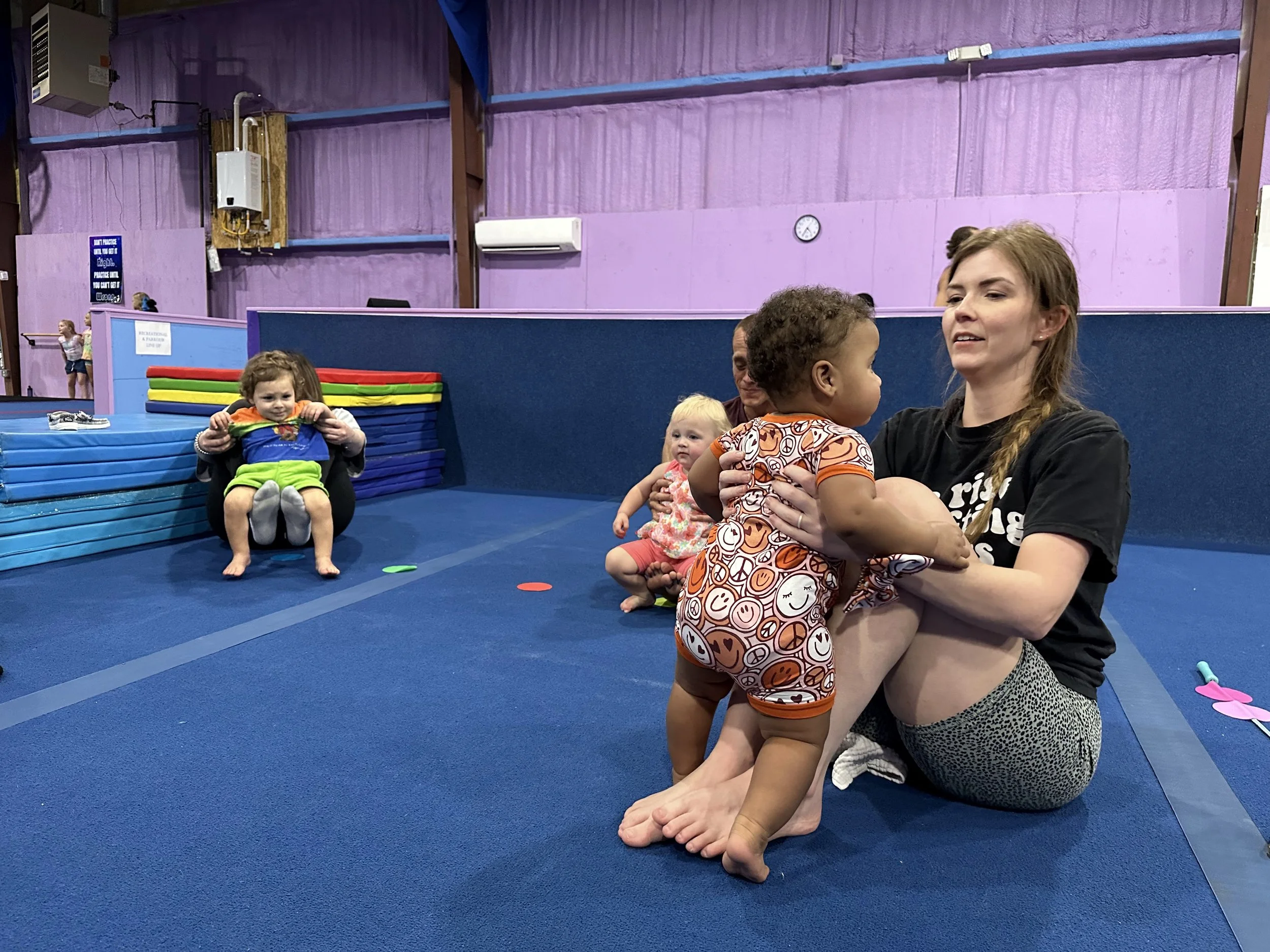 Group of children and a woman in a gymnasium with purple walls and blue mats, some children sitting on the mats and one child being held by the woman.