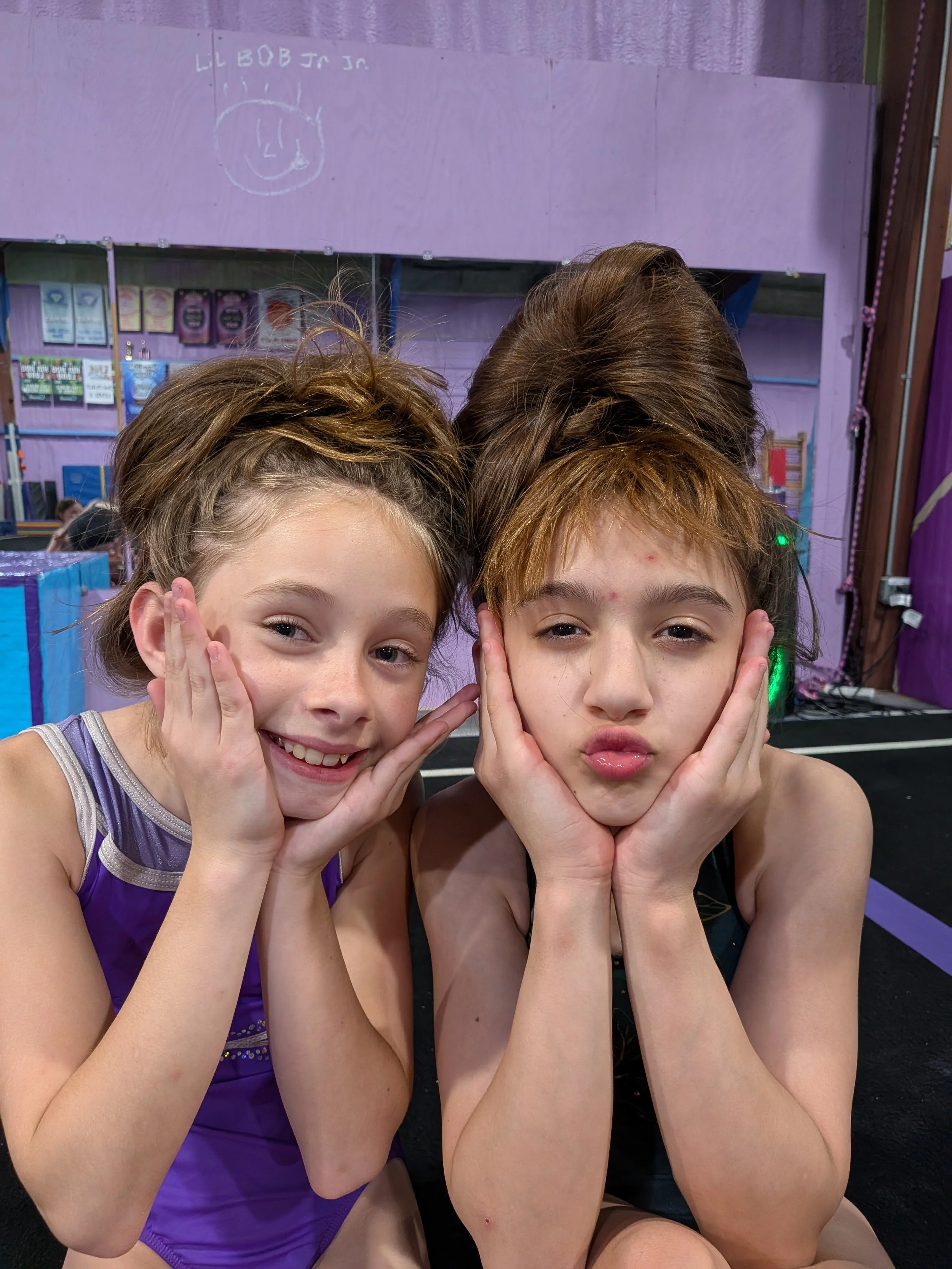 Two young girls with curly hair posing with their hands on their cheeks, smiling and making similar face expressions, sitting close together in a gymnastics or dance studio.