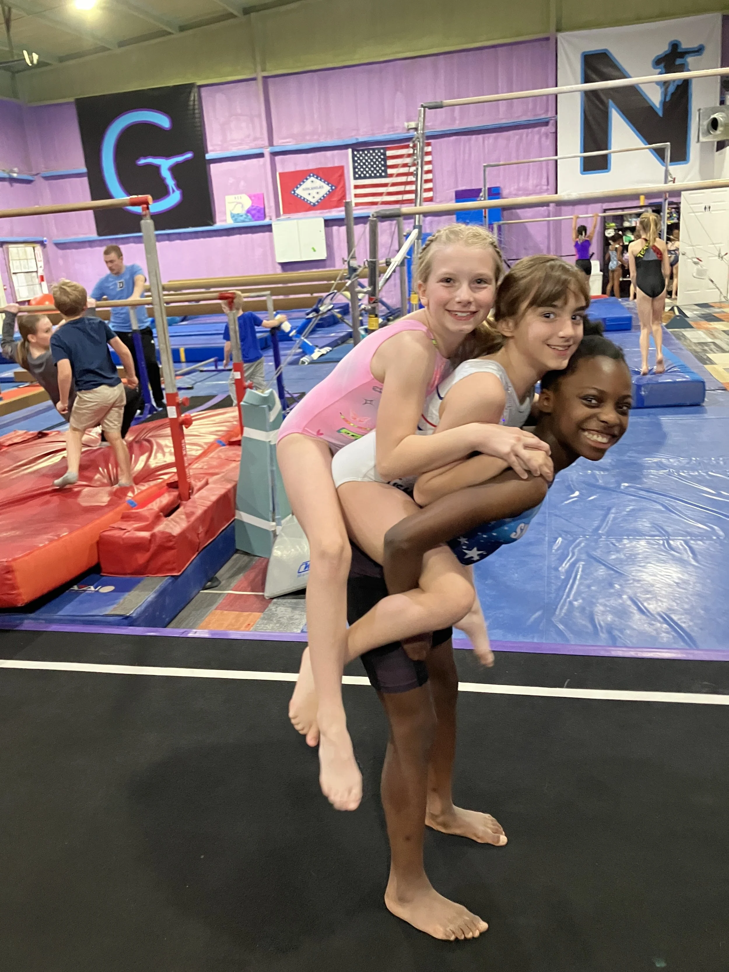 Three young girls, one with blonde hair, one with light brown hair, and one with dark skin and black hair, are practicing acro yoga or gymnastics in a gymnasium, with one girl standing on the shoulders of another, smiling at the camera. The background shows a gymnastics area with mats, balance beams, and other children and adults.