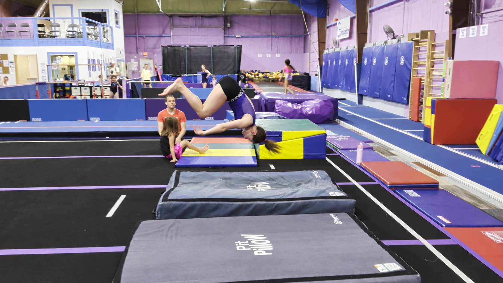 Two children practicing gymnastic moves on a blue mat inside a gymnasium. The girl is in a plank position with her arms extended and legs raised, while the boy is supporting her from underneath. Hot Springs Gymnastics | 112 Bluestone Ct. Hot Springs,