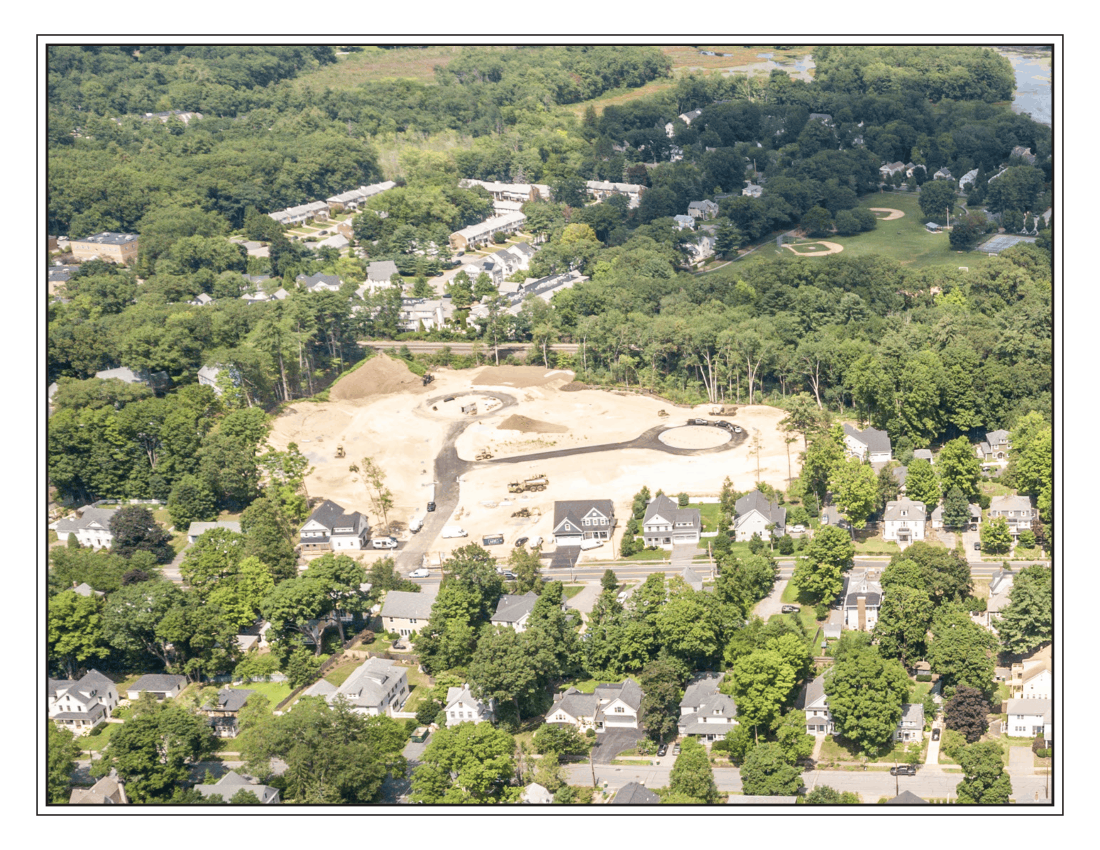 Aerial view of a suburban neighborhood with numerous houses surrounded by trees, a park with baseball fields, and a construction site in the foreground.