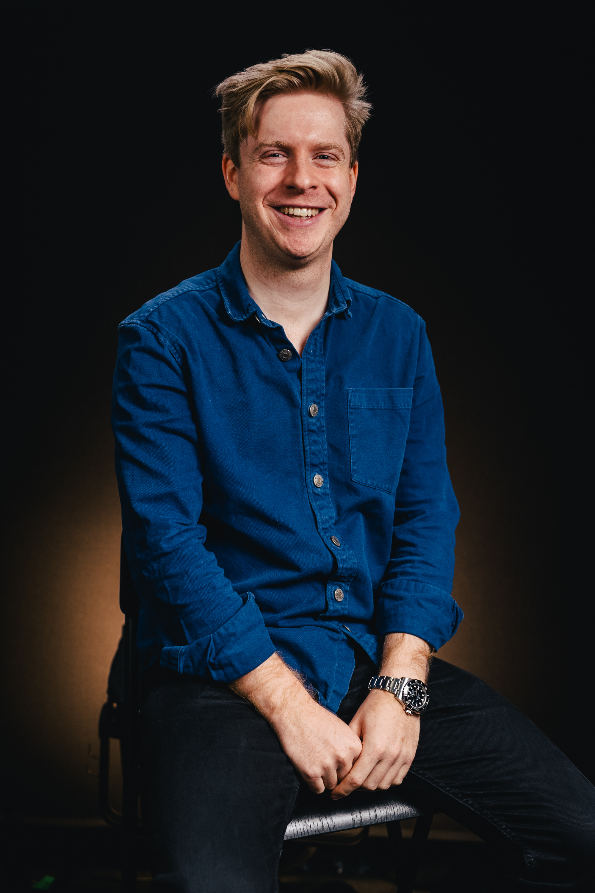 A young man with blonde hair and blue eyes smiling, wearing a blue button-up shirt, sitting on a chair with a dark background.