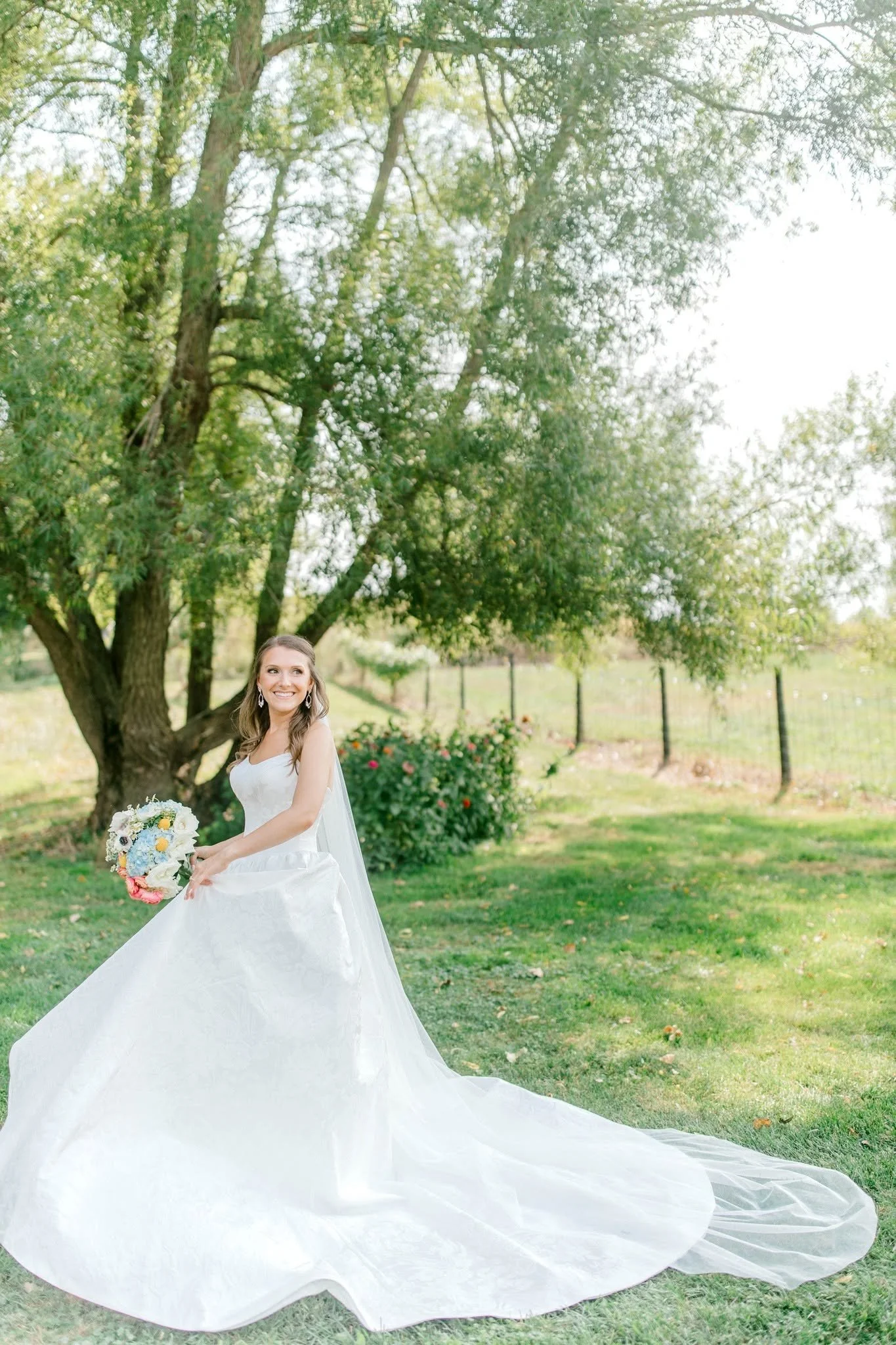 A bride in a white wedding gown holding a bouquet of flowers outdoors next to a large tree with green leaves.