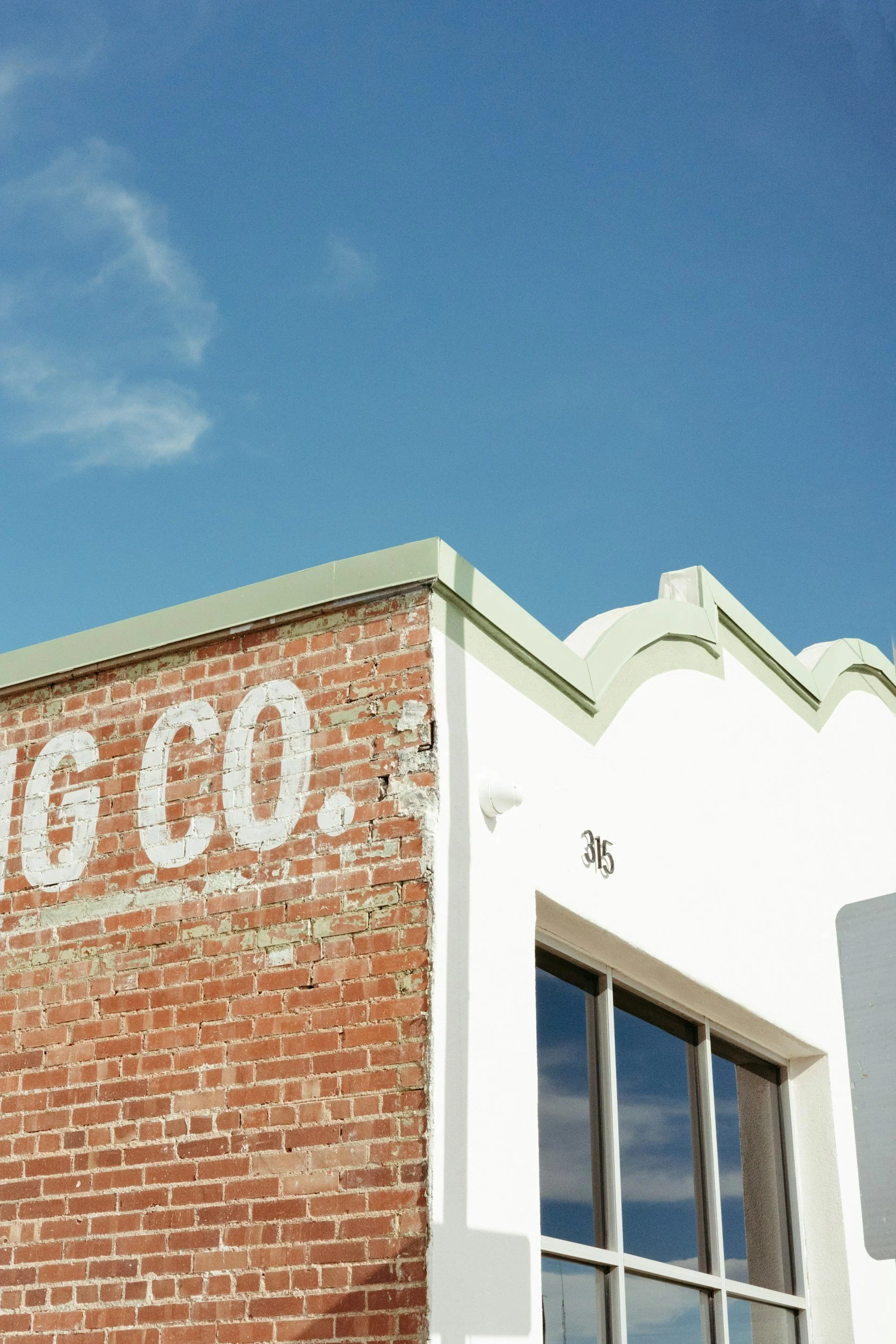 Part of a building with a red brick wall, large white text painted on the brick, white stucco, and a window with reflective glass under a clear blue sky.