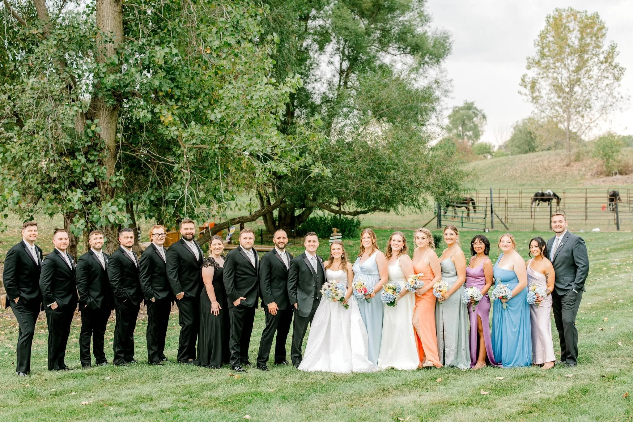 A group of people dressed in formal attire, standing outdoors in a park-like setting with green grass and trees in the background, during daytime.