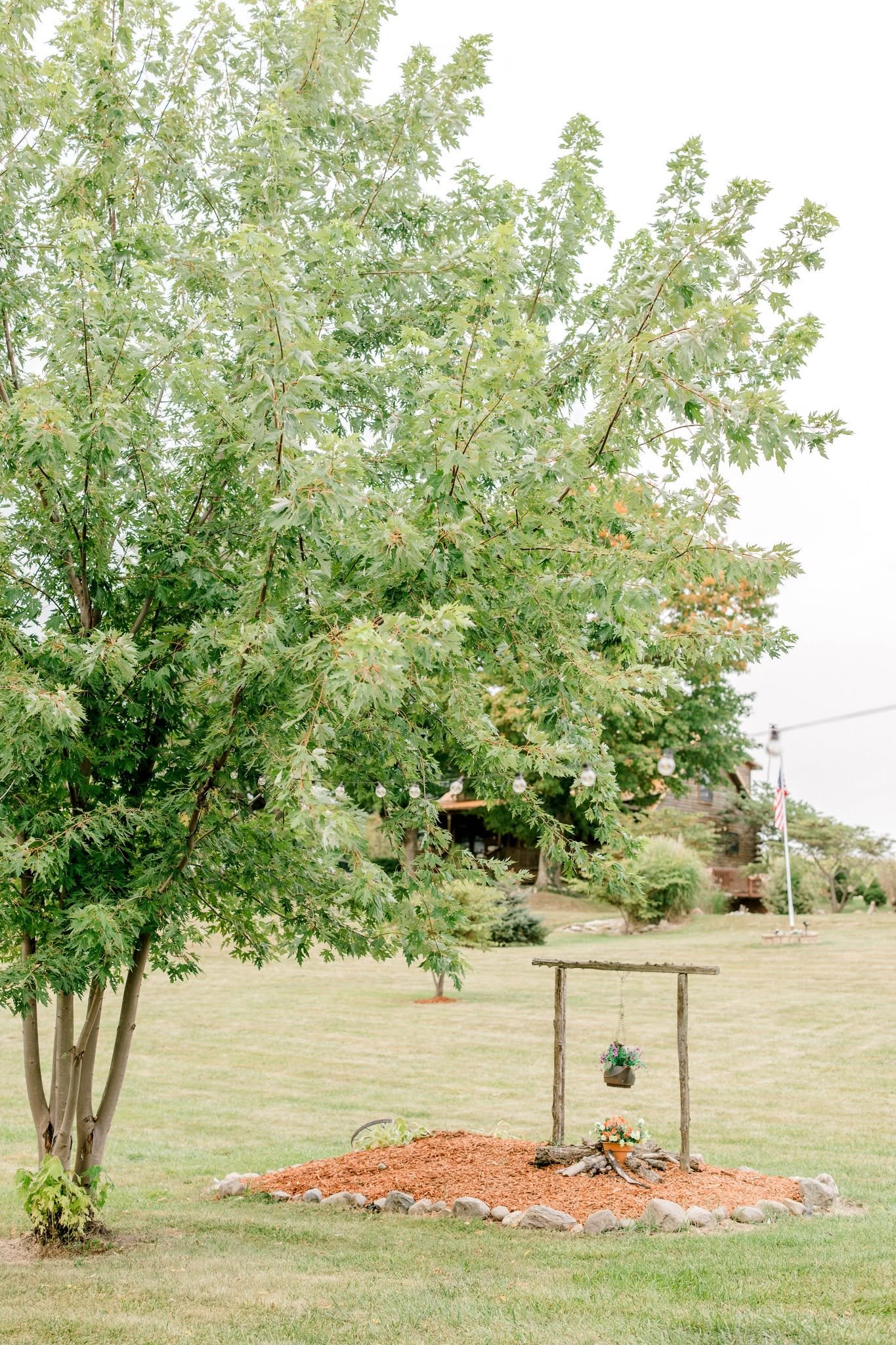 A grassy yard with a large tree on the left, a small wooden bench and a decorated flower garden area in the center, and a house with an American flag in the background. There are string lights hanging in the yard.