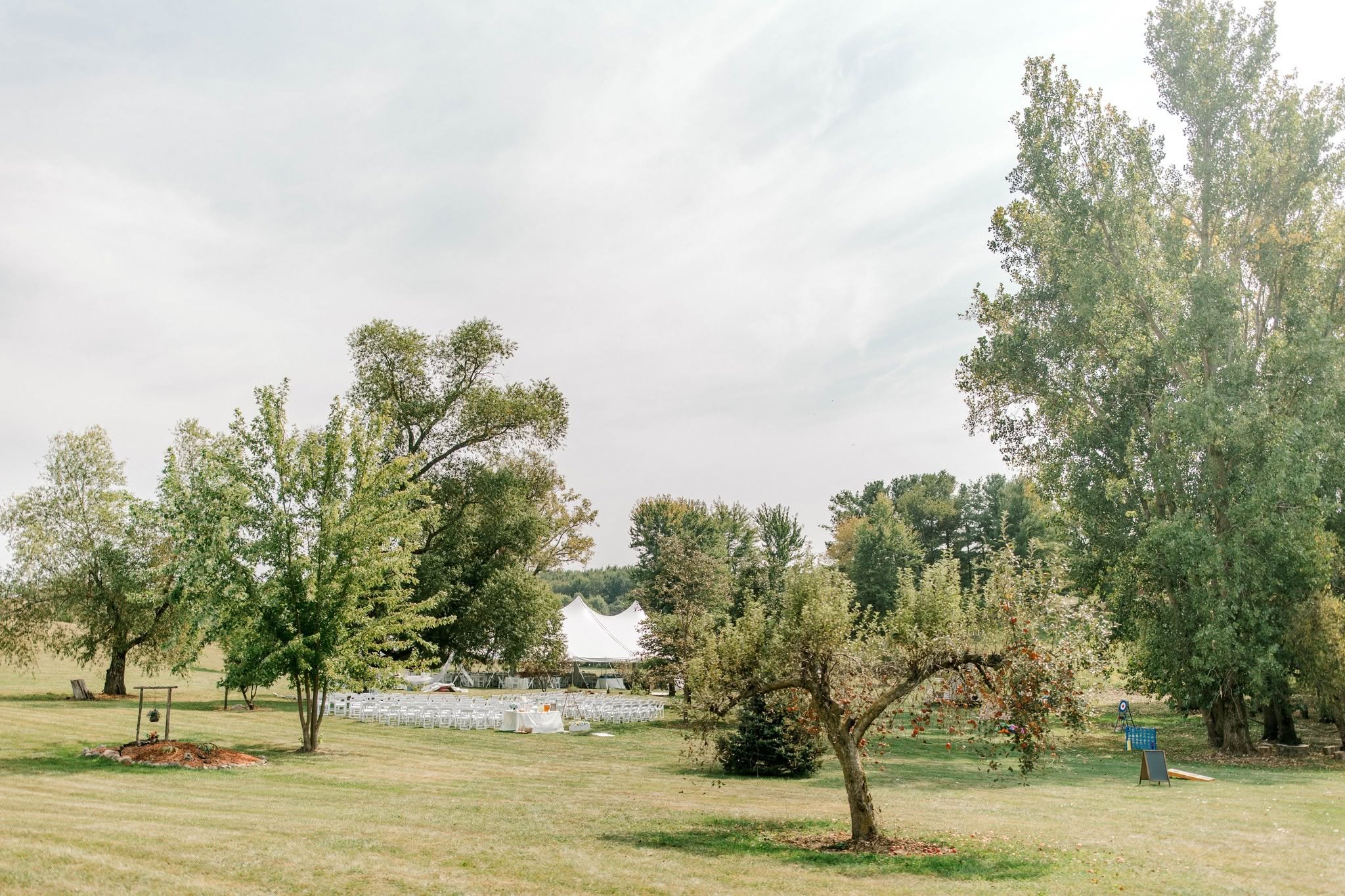 An outdoor event setup in a park with white chairs arranged in rows, surrounded by lush green trees and a white event tent in the background. The sky is partly cloudy.