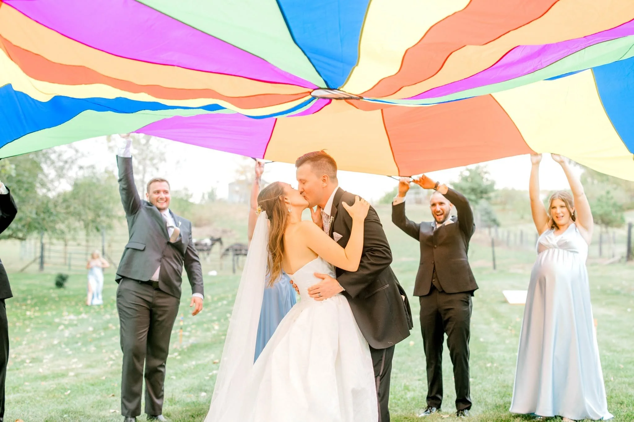 A bride and groom kiss under a large, colorful, rainbow-like parachute with several people holding it up around them, outdoors in a park-like setting.