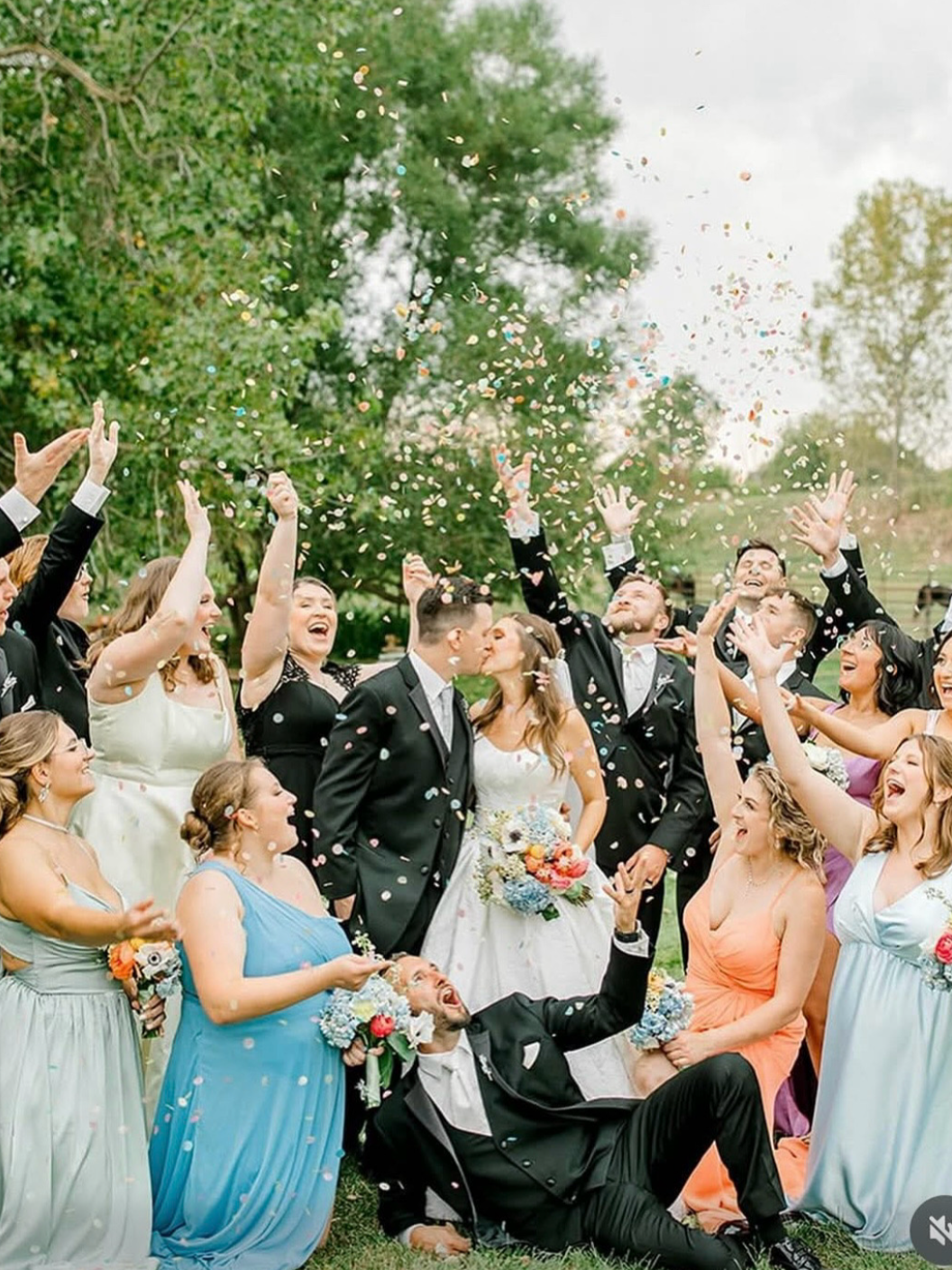 A group of people at a wedding celebration outdoors, throwing confetti and celebrating, with a bride and groom in the center sharing a kiss.