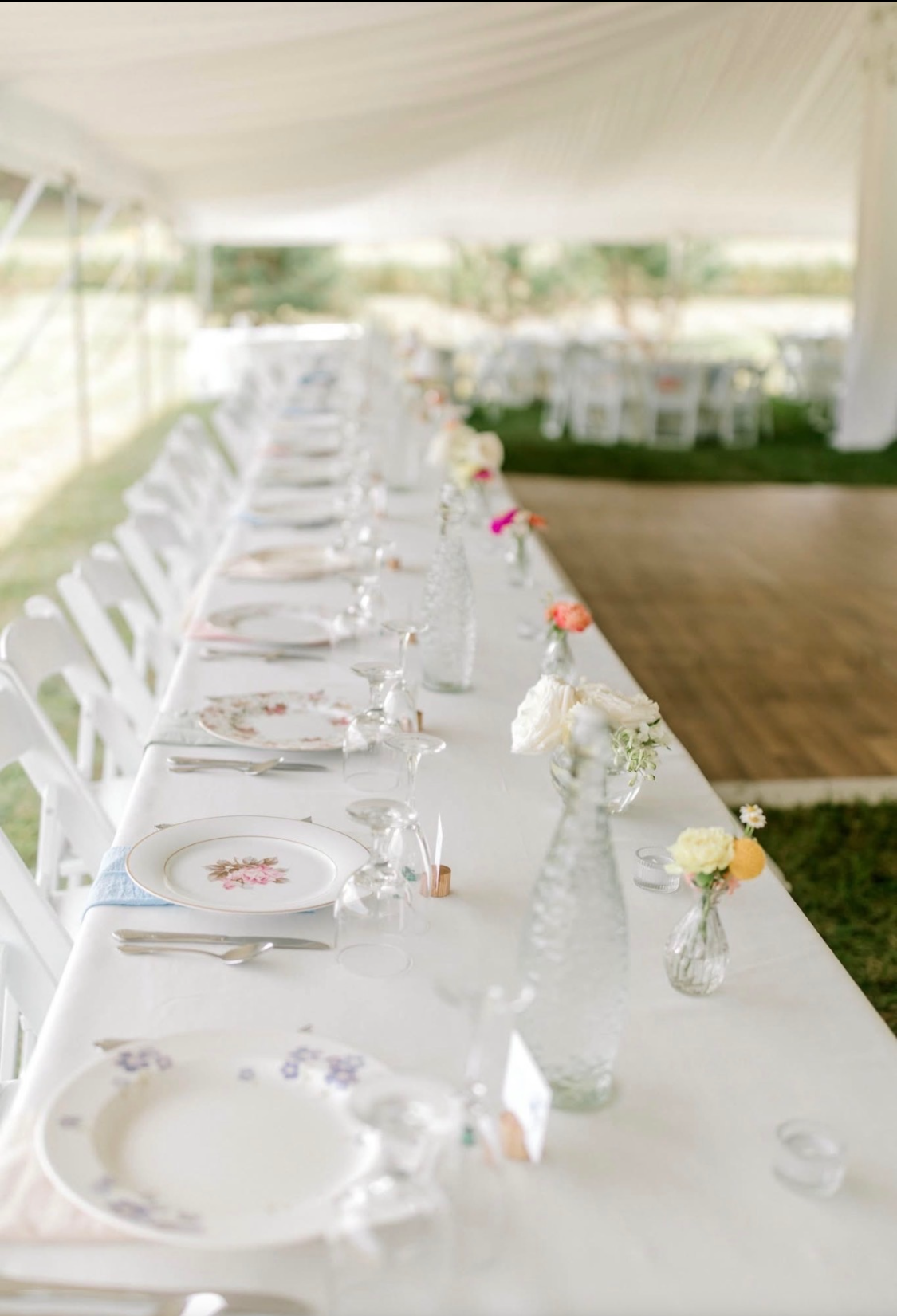 Long banquet table set for a wedding or special event, decorated with white tablecloth, floral centerpieces in small glass vases, and place settings with floral plates, silverware, and glassware under a tent with green grass and trees outside.