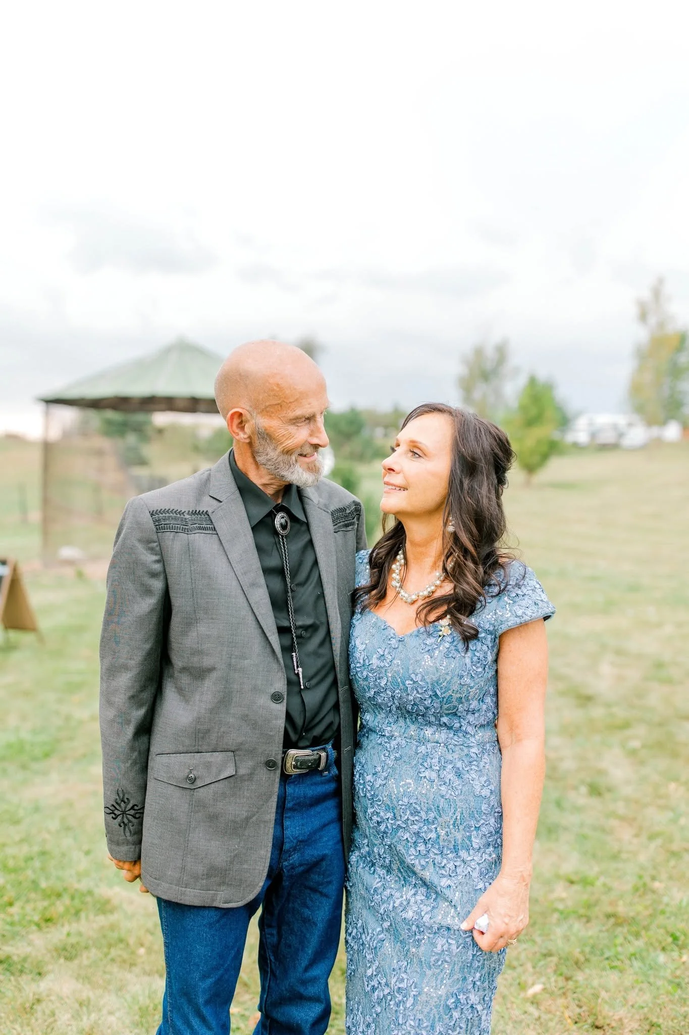 An older man and woman standing outdoors on grass, looking at each other and smiling, with trees and a small pavilion in the background.