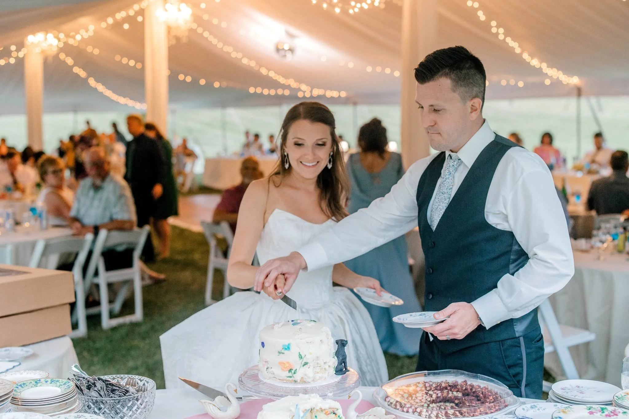 A bride and groom cutting a wedding cake at their reception under a tent decorated with string lights, with guests seated at tables in the background.
