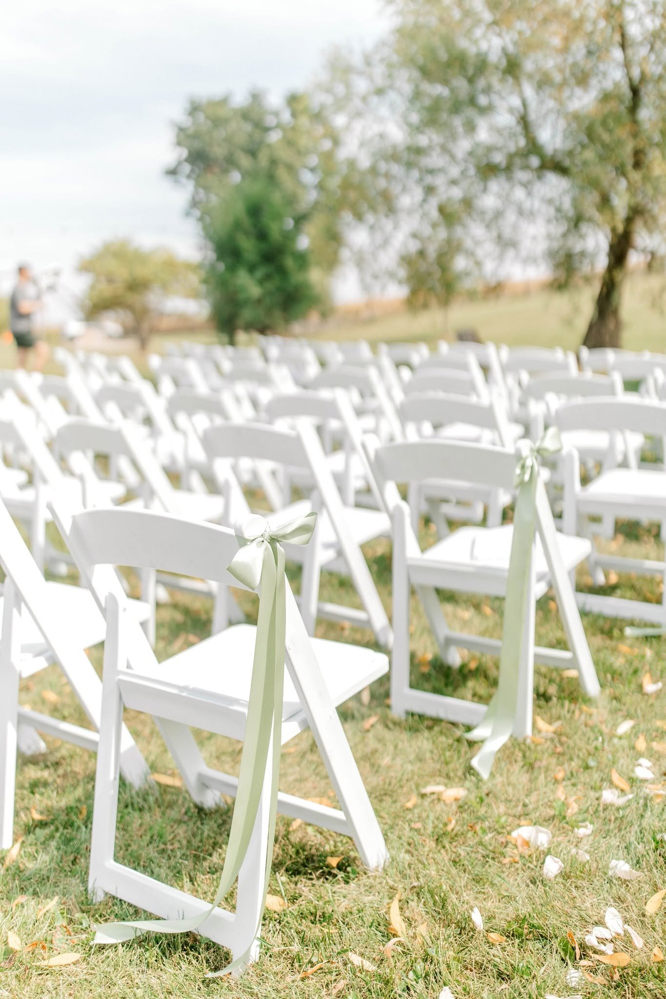 Empty white folding chairs with cream ribbons tied on the sides, set up outdoors on grass with trees in the background for a wedding ceremony.