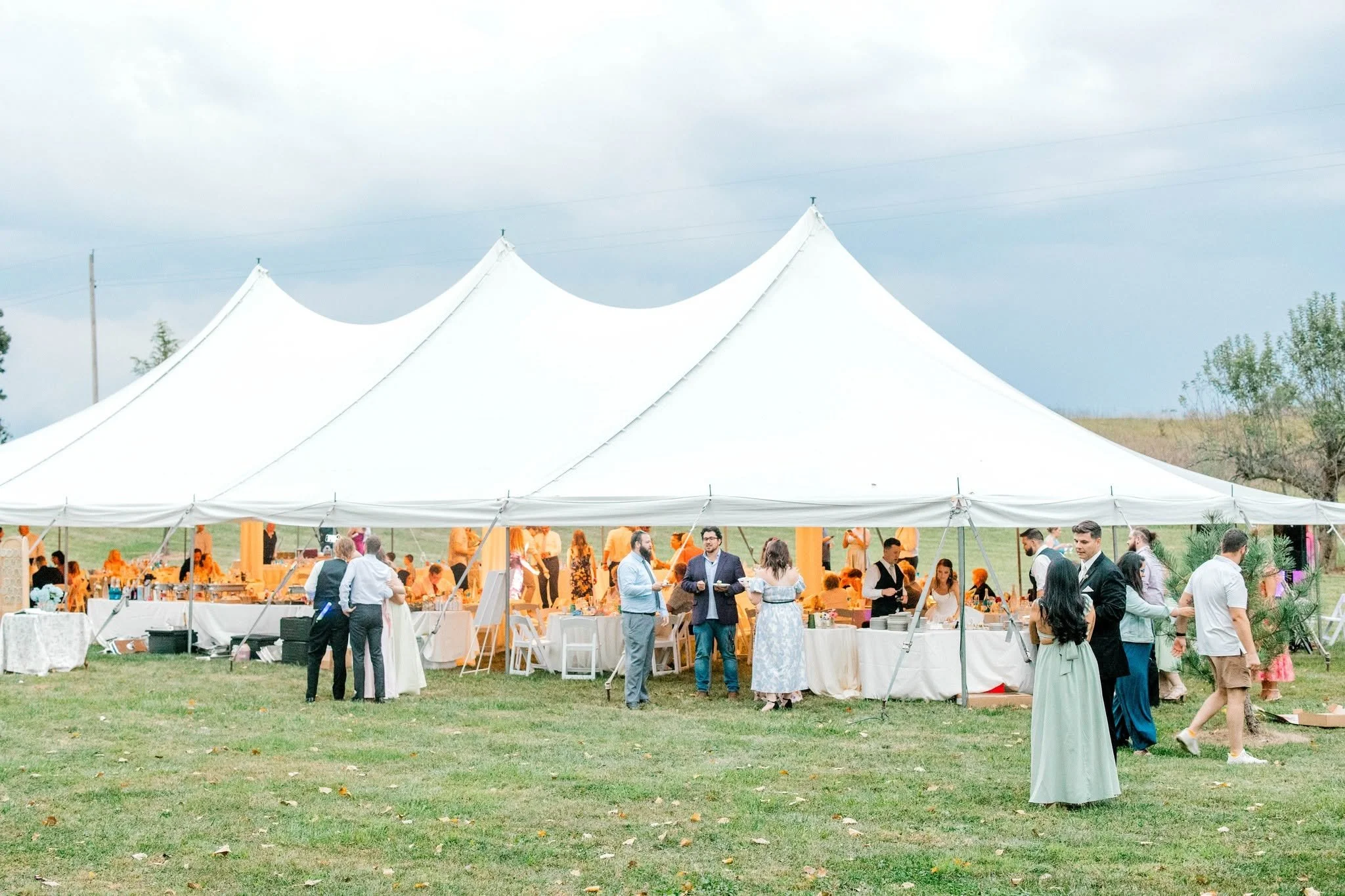 People gathering at an outdoor event under a large white tent on a grassy field, with tables and food.