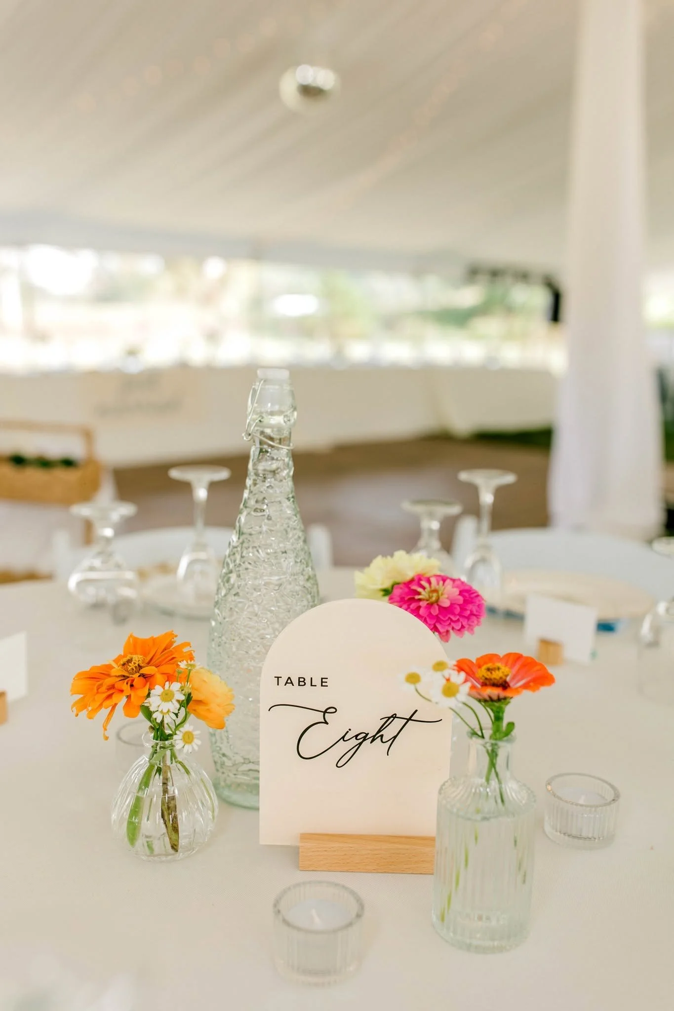 Table decor with small glass vases filled with colorful flowers, a bottle of water, and a sign that reads 'Table Eighth' in cursive printing.