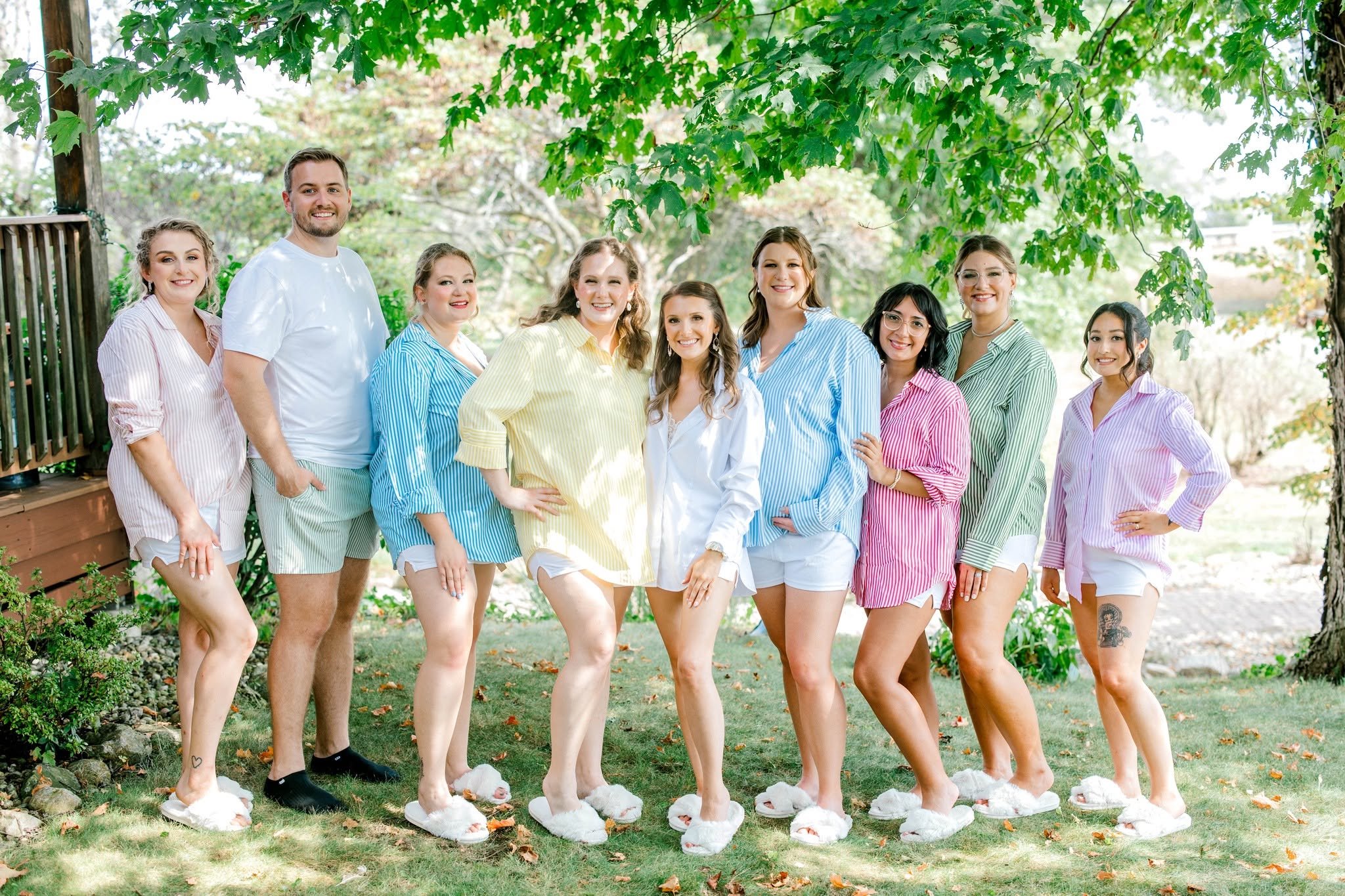 A group of ten people, mostly women and one man, standing outdoors under a large green leafy tree, wearing pastel-colored striped pajamas and fluffy slippers, smiling at the camera.