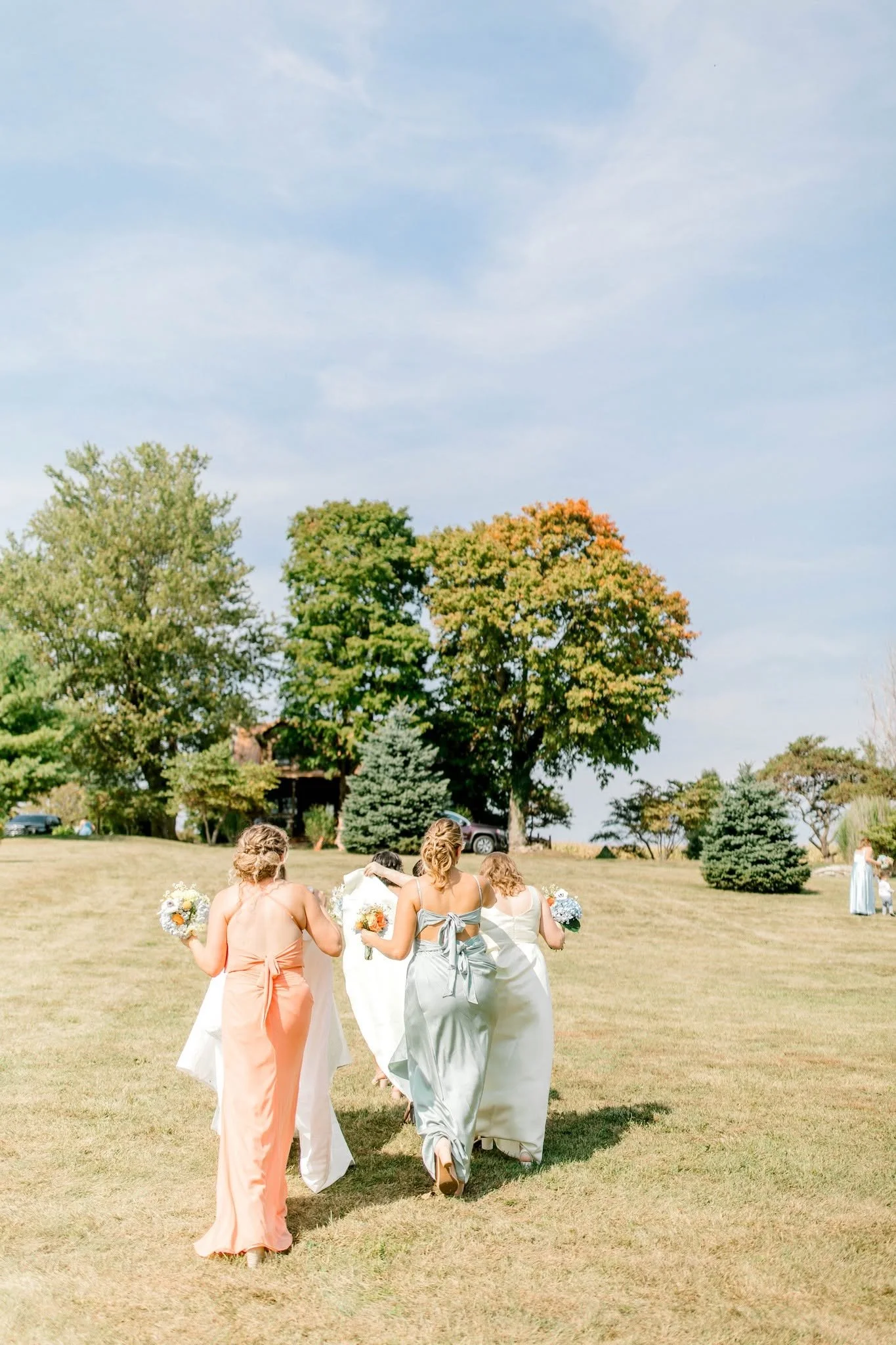 A group of women in dresses walking on a grassy field carrying bouquets, with trees and a clear sky in the background.