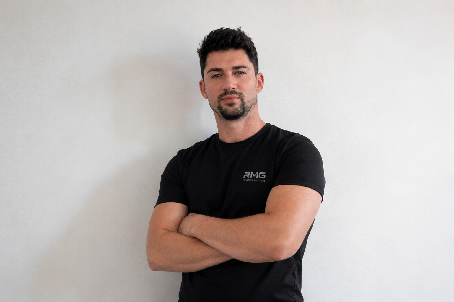 Hombre joven con barba y cabello oscuro, usando camiseta negra con logo en el pecho, posando con brazos cruzados en una pared blanca.