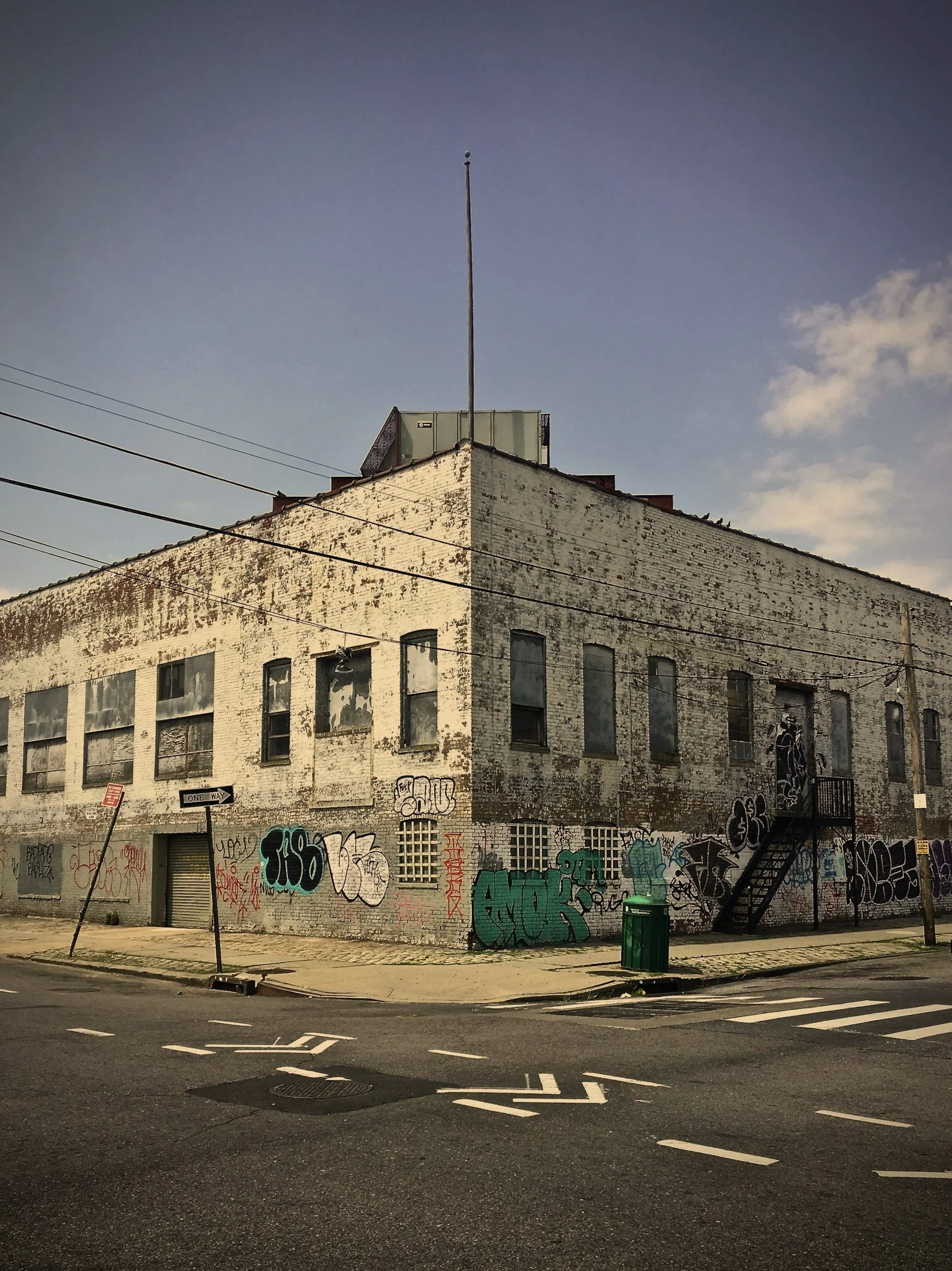 Sycamore Street Advisors, worn brick building with multiple windows, graffiti on the walls, and a metal staircase outside. The building is located on a street corner with a one-way sign, a green trash bin, and power lines overhead.