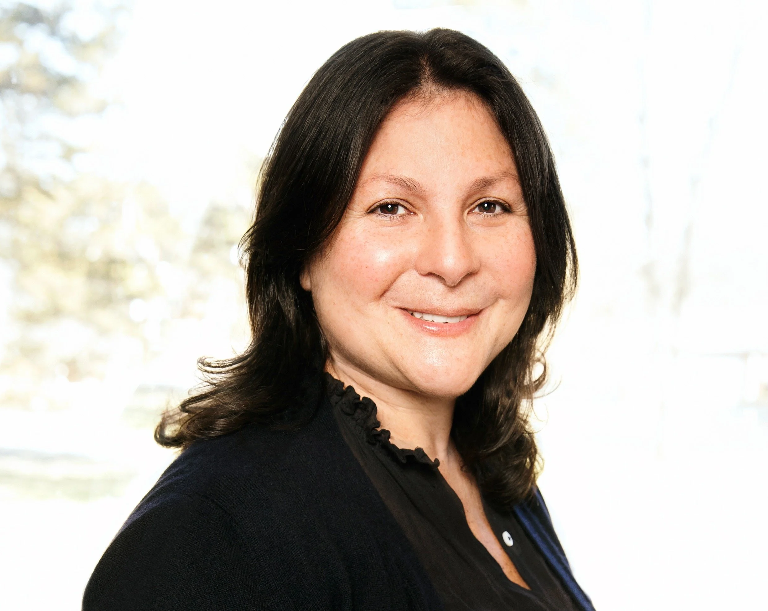 Samara Daly, of Sycamore Street Advisors picture with dark hair and light skin smiling, wearing a black top, with a blurred outdoor background.