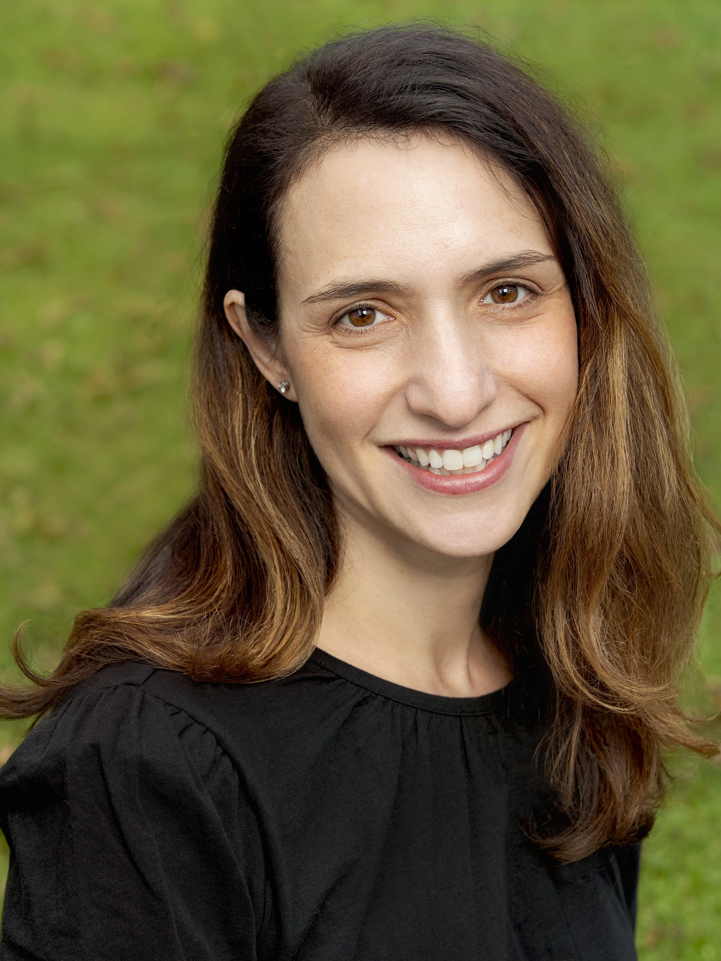 Eloise Paul of Sycamore Street Advisors  brown hair and hazel eyes smiling outdoors, wearing a black top, with a blurred green background.