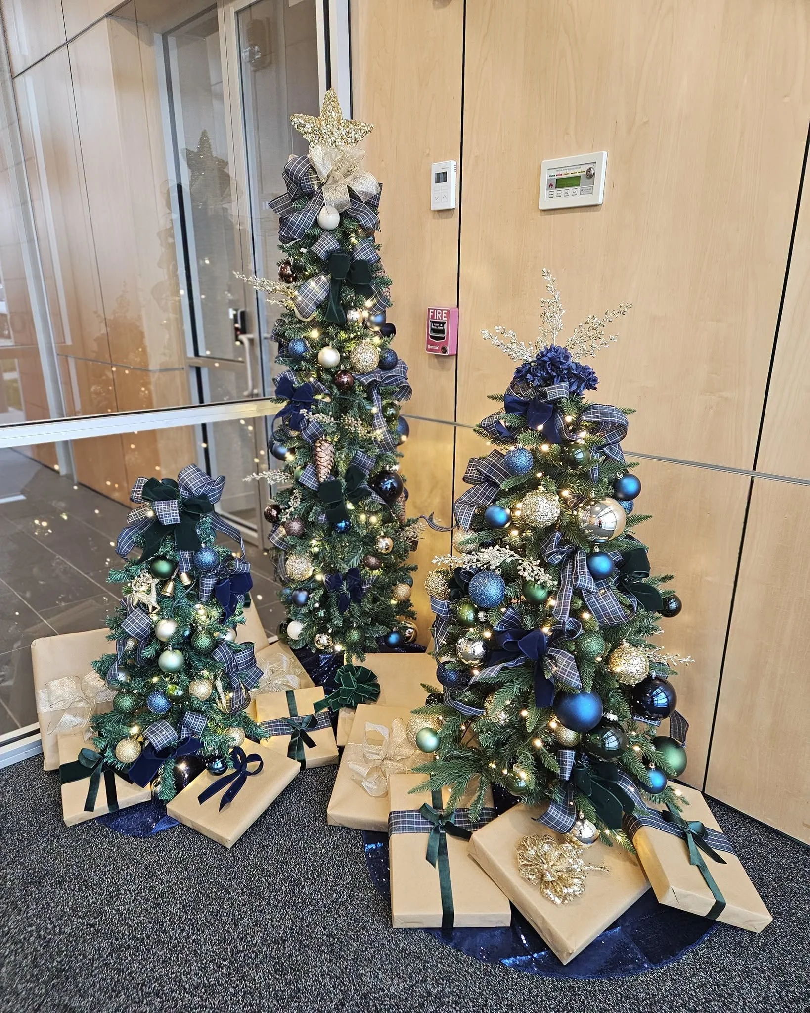 Three decorated Christmas trees with ornaments, ribbons, and a gold star topper, surrounded by wrapped presents on a carpeted floor in front of a wooden wall with a fire alarm and thermostat.