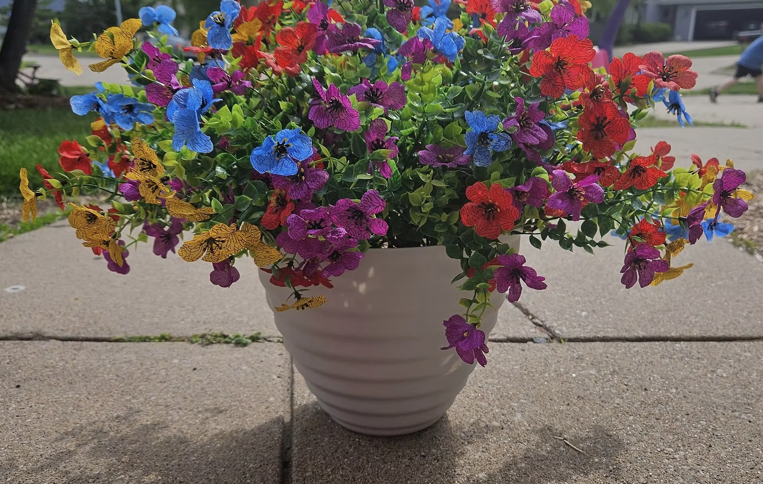Colorful pot of blooming petunias with purple, red, yellow, and blue flowers on a sidewalk.