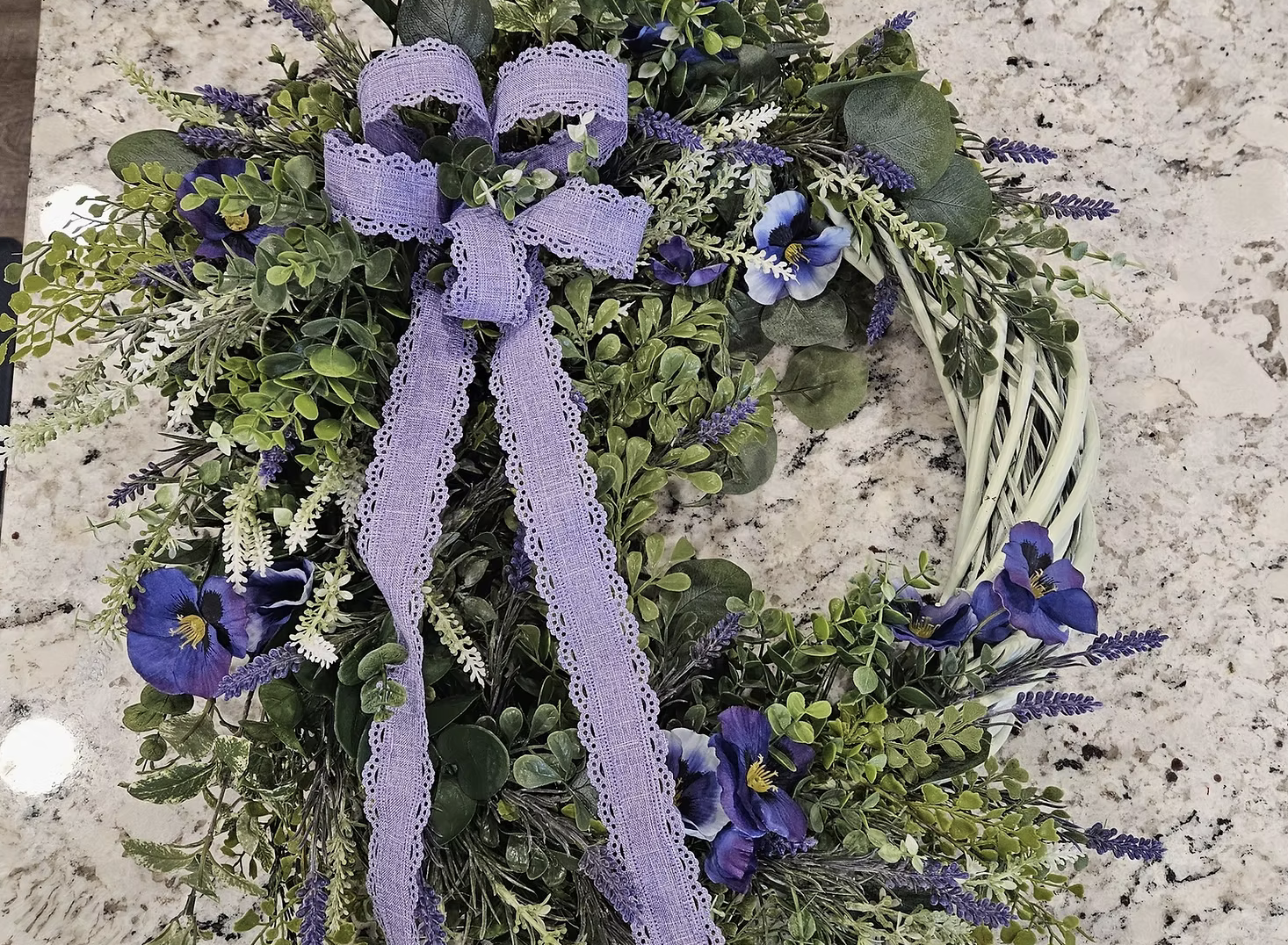 A decorative wreath made of green foliage and purple flowers, tied with a purple lace ribbon, placed on a granite countertop.