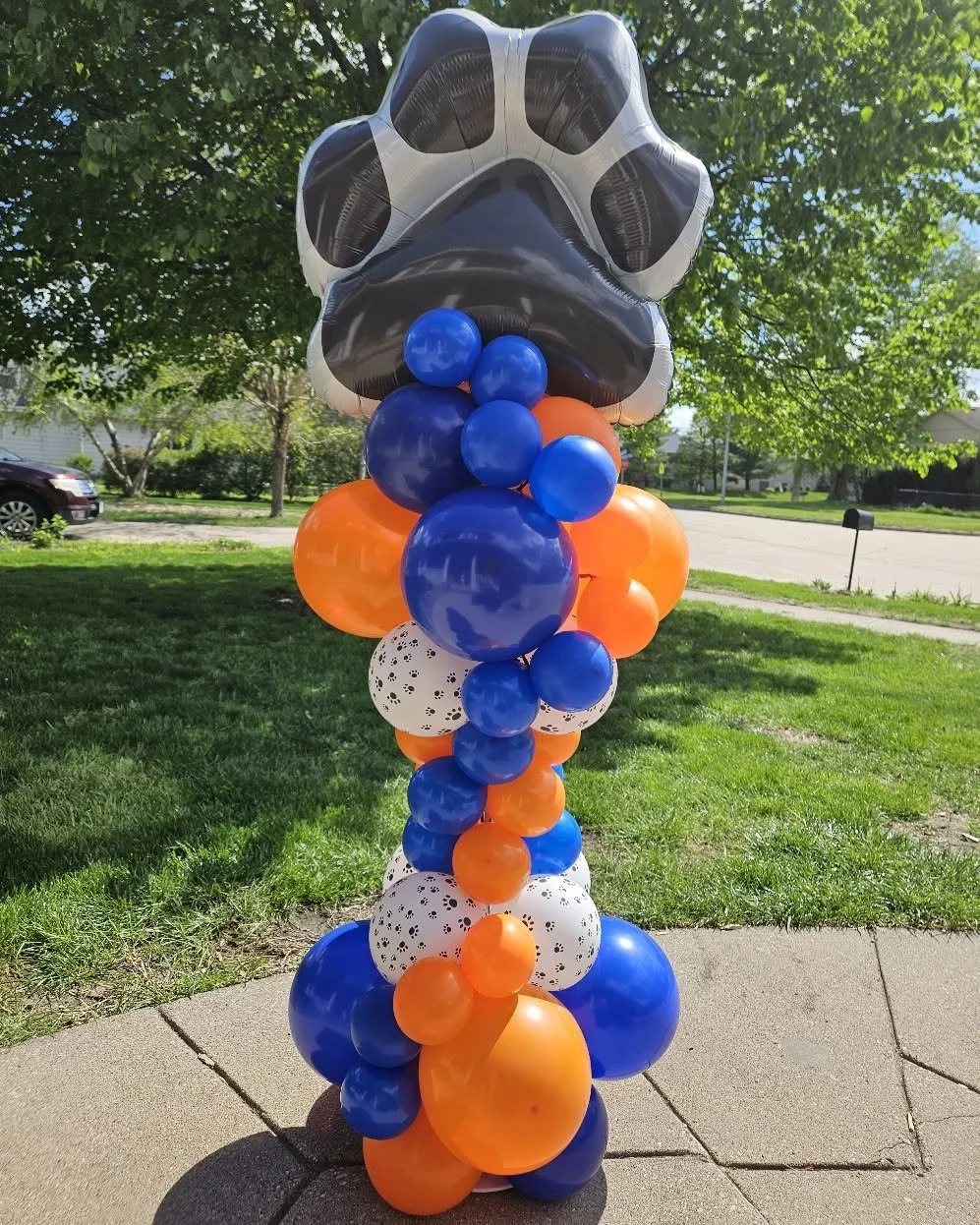 Balloon column with a large animal paw print balloon at the top, surrounded by orange, blue, and white balloons, set outdoors on a sidewalk with grass and trees in the background.