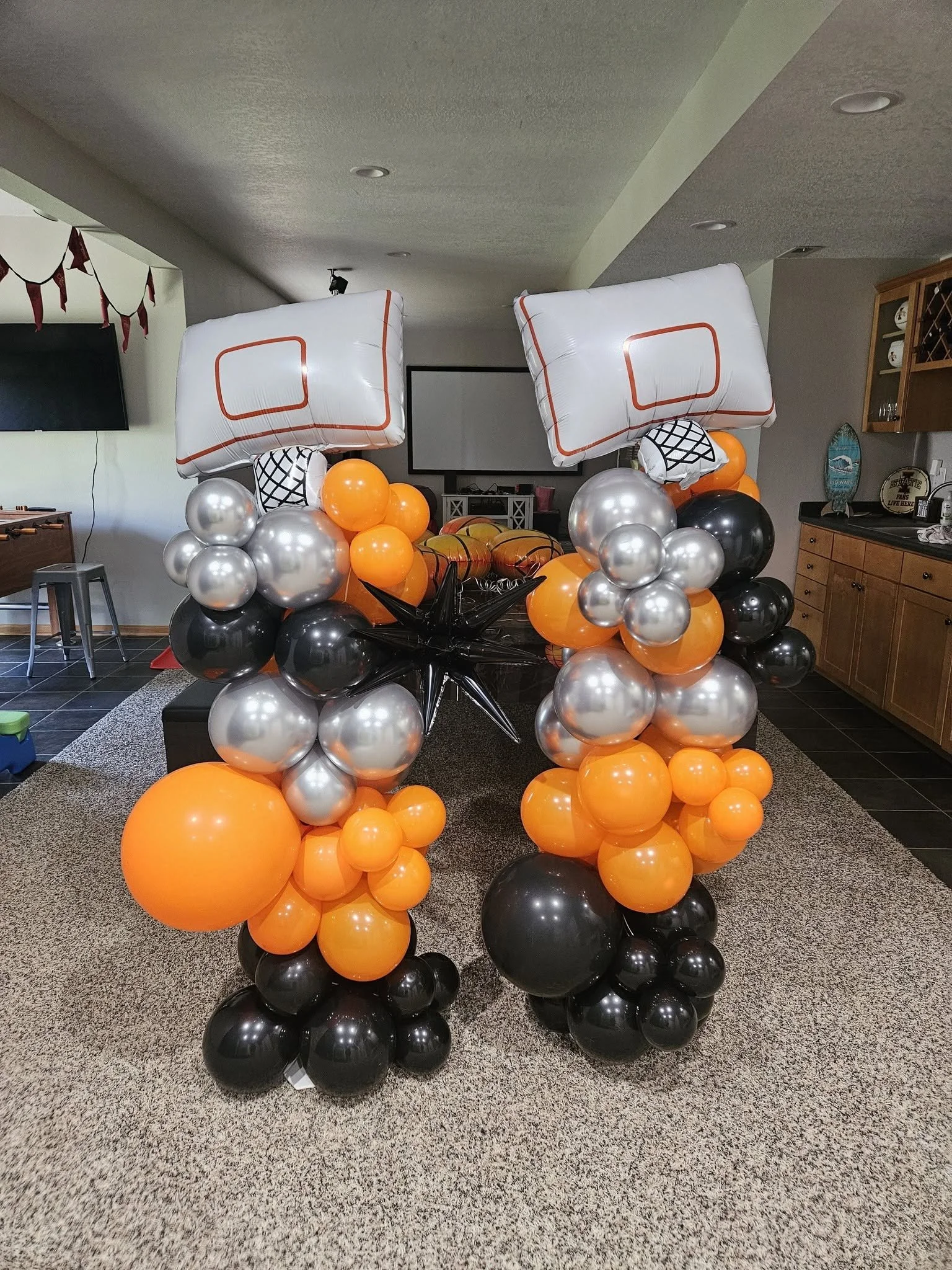 Decorative balloon columns with orange, black, and silver balloons, topped with large white balloons displaying basketball court lines, inside a living room.