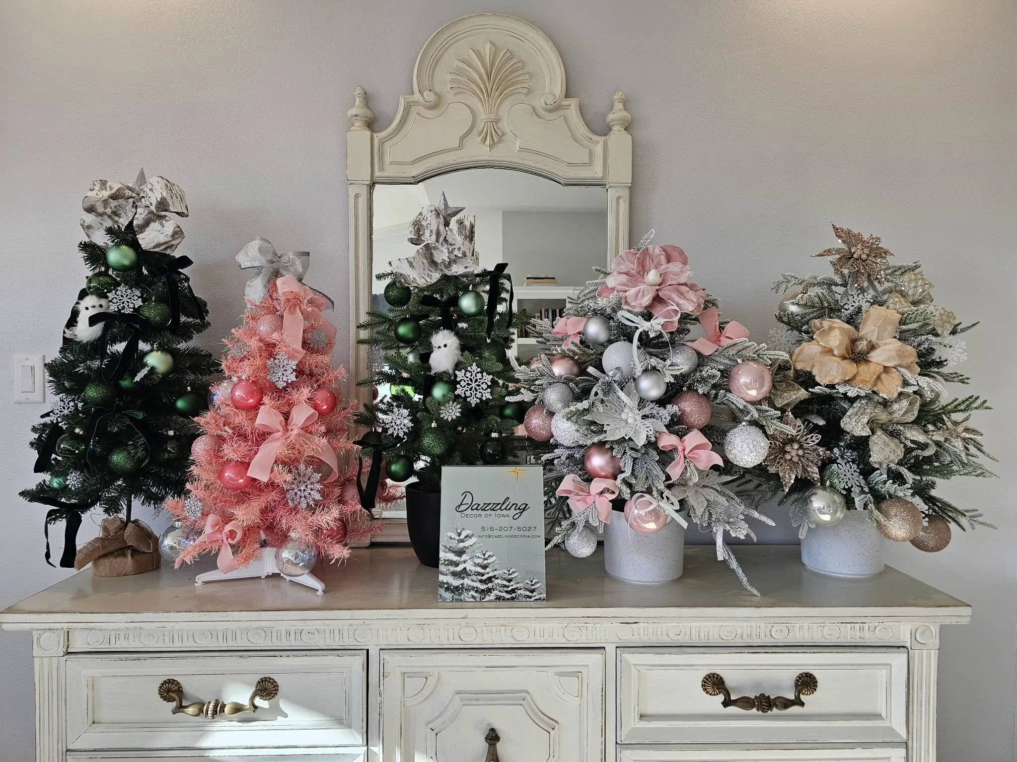Four decorated Christmas trees in various colors and styles are arranged in front of a large mirror on a white wooden sideboard. The trees are adorned with ornaments, ribbons, and decorative items in pink, green, silver, and gold. A sign on the sideboard reads ‘Dazzling Decor of Iowa’ with contact information.