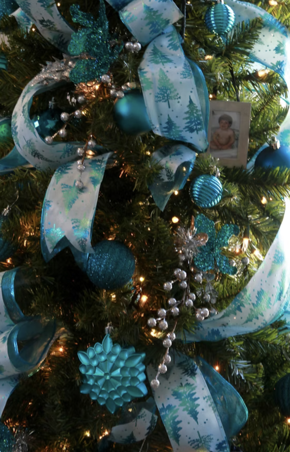 Close-up of a decorated Christmas tree with blue and silver ornaments, ribbons, a small photo, and string lights.