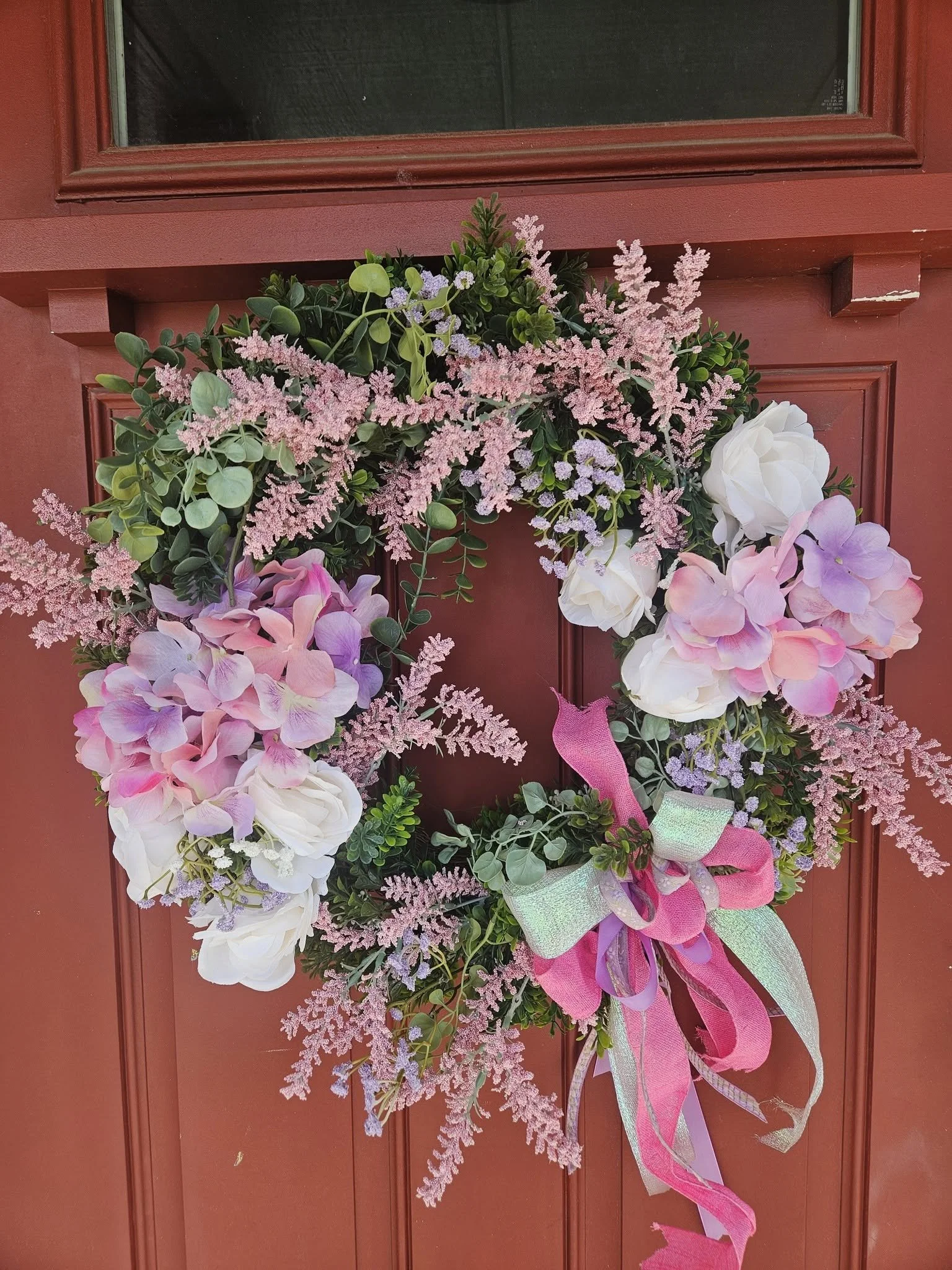A door decorated with a floral wreath made of pink, purple, and white flowers, and green foliage, with pink and green ribbons hanging from it.