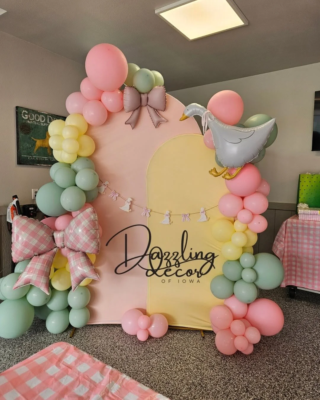 Decorative display with pastel-colored balloons, pink and green, arranged around a pink backdrop featuring a yellow half-circle. The display includes a large pink and white checkered bow, a paper banner with small white dresses and pink bows, and a gray inflatable stork. The bottom of the image has pink and white checkered tablecloths. The display promotes Dazzling Decor of Iowa.