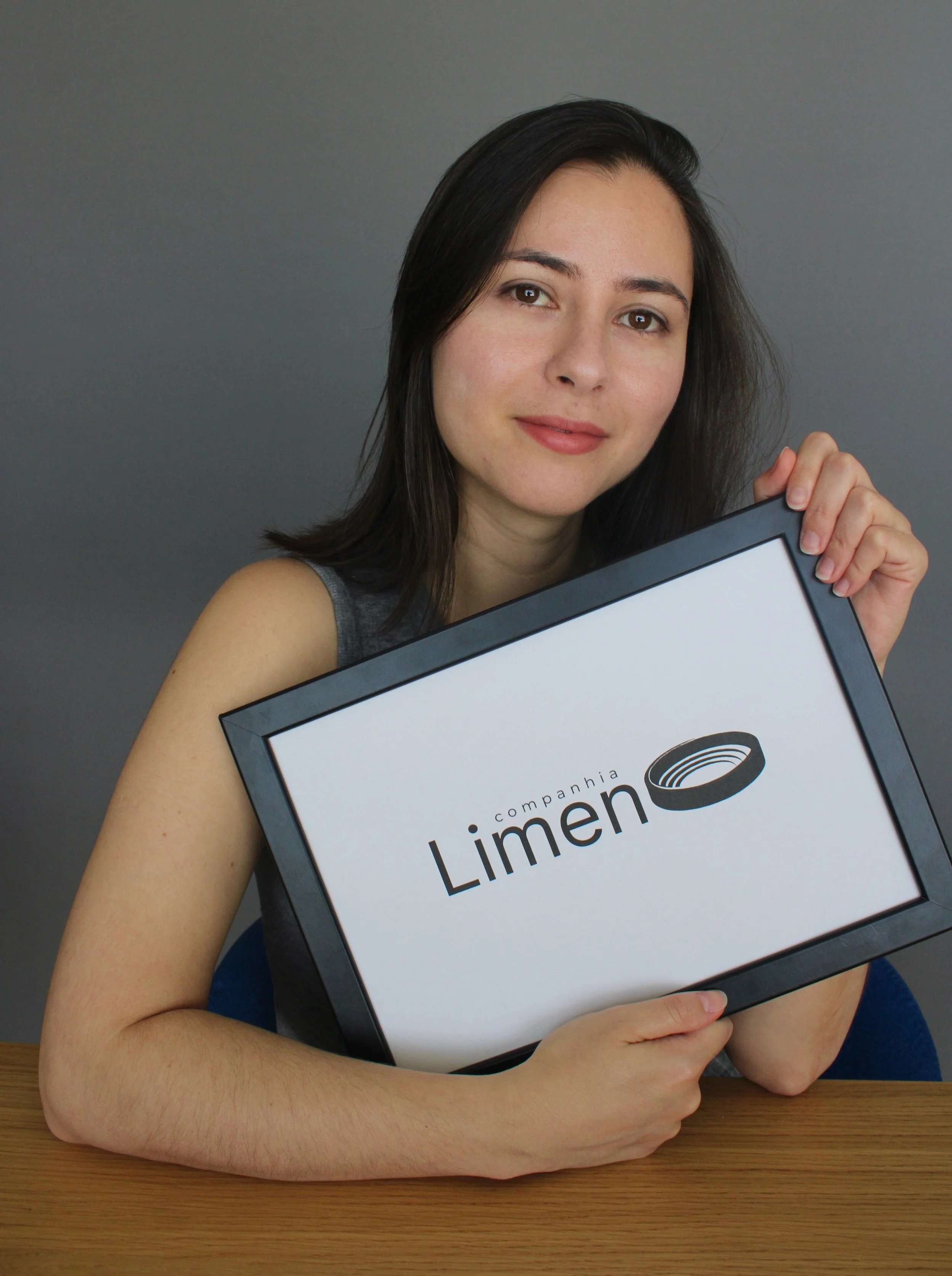 A cofundadora da associação, Thaís Chanes, segurando uma placa com o logo "Companhia Limen" em um fundo cinza, apoiada em uma mesa de madeira.