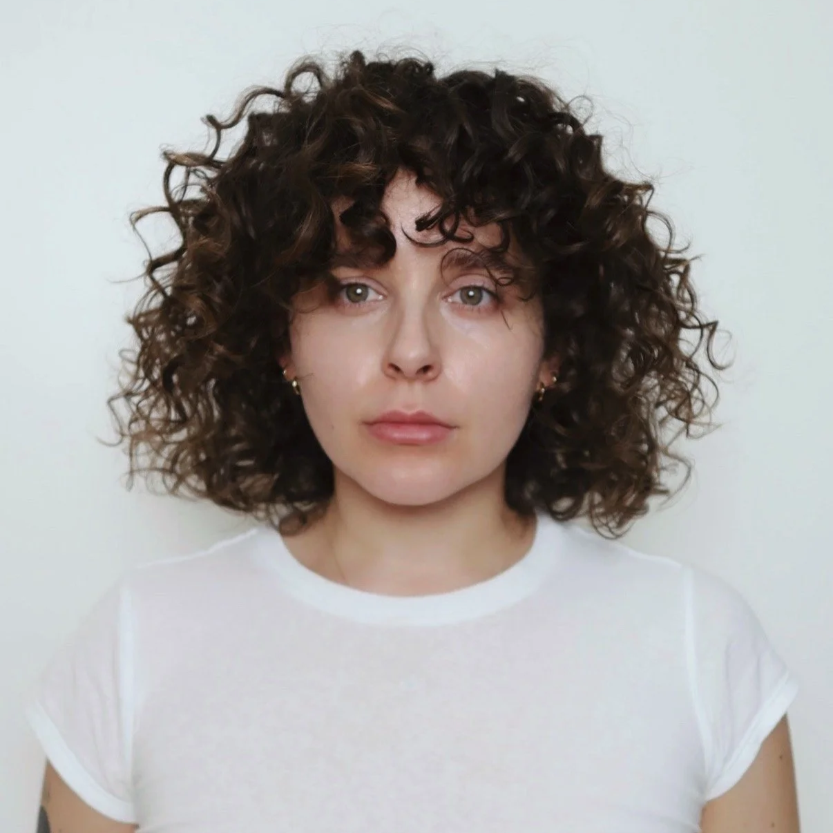 A woman with curly brown hair and light skin, wearing a white t-shirt and small earrings, looking directly at the camera against a plain white background.