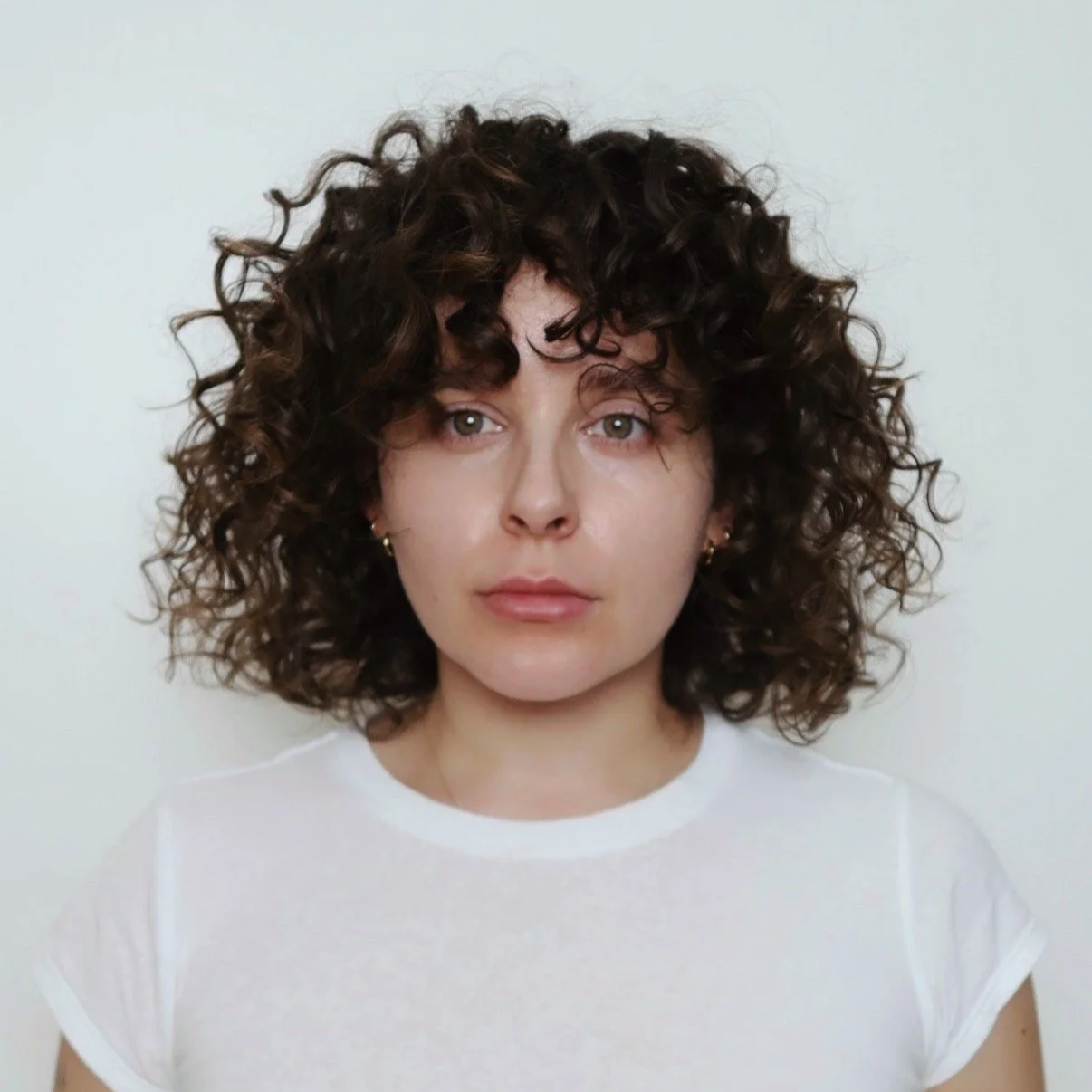 Close-up portrait of a woman with short, curly dark hair, light skin, and wearing a white T-shirt, against a plain light background.