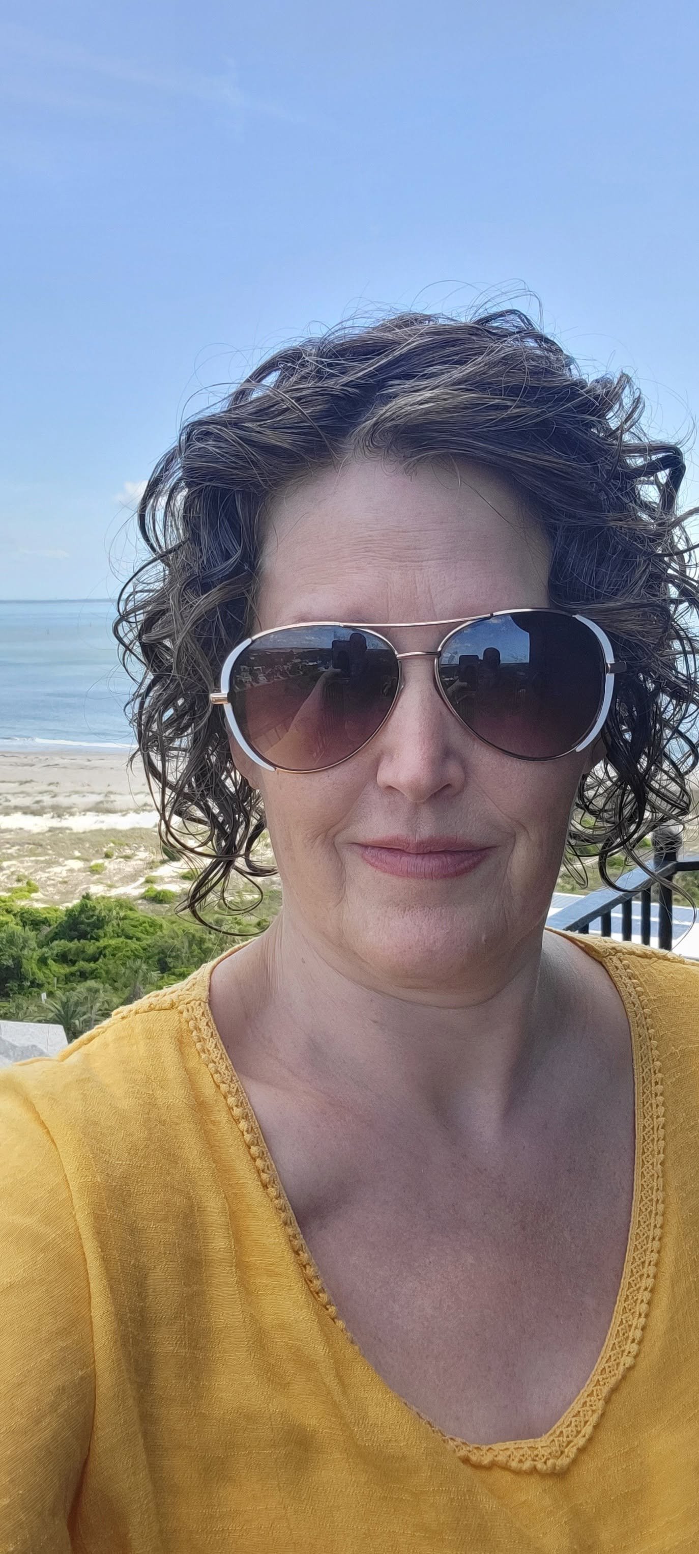 A woman with curly hair wearing sunglasses and a yellow top standing on a beach with the ocean in the background.