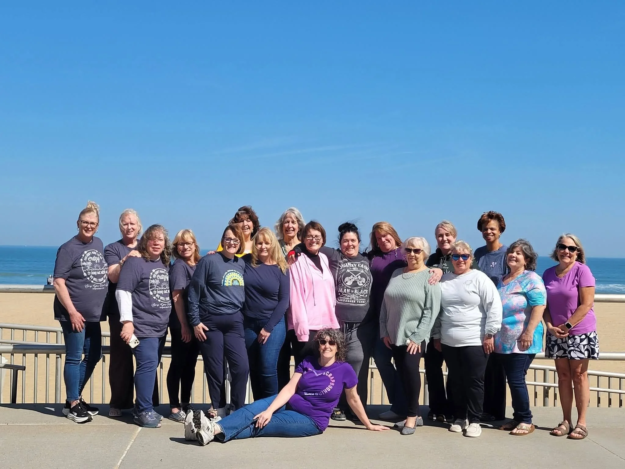 Group of women and one young girl posing on a boardwalk near the beach with blue sky and ocean in the background.