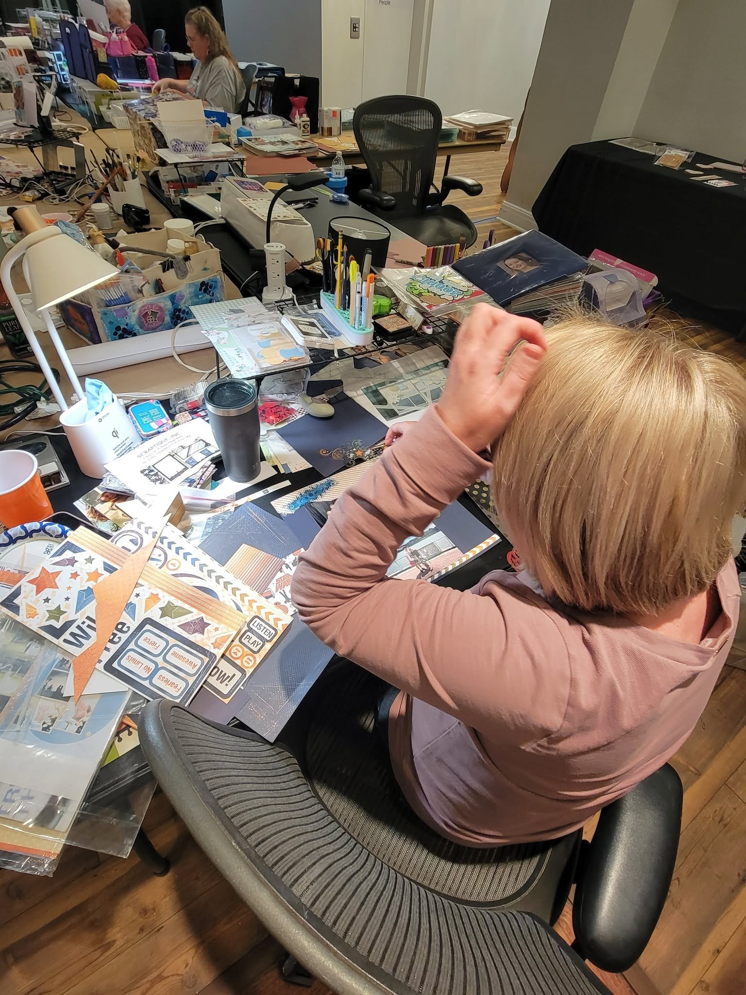 A person with blonde hair and a pink jacket sitting at a cluttered table with various papers, office supplies, and craft materials, in a room with other people working at desks.
